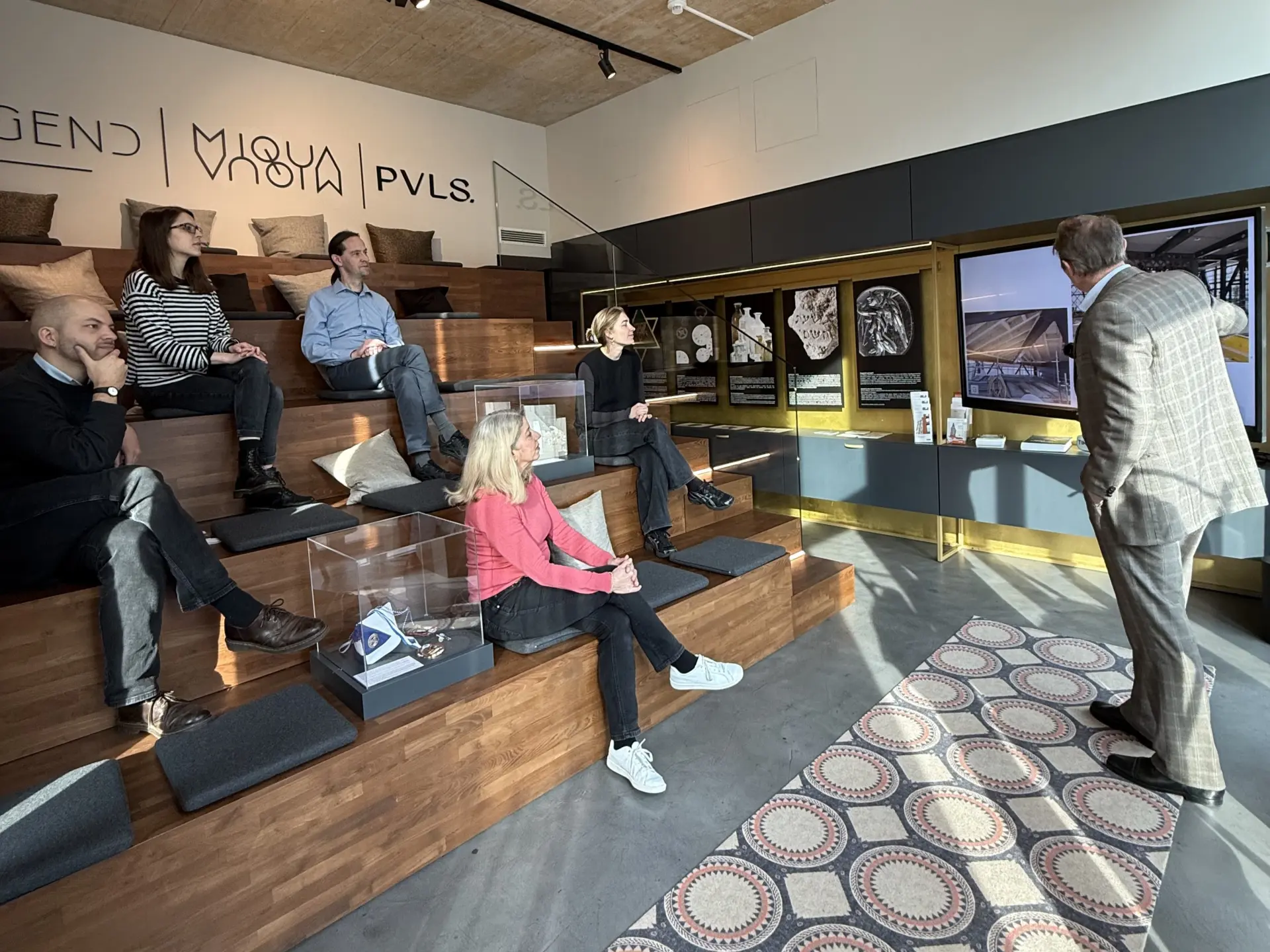 Eine Gruppe Menschen sitzen auf einer Treppe in einem Raum und hören einem Vortragenden zu.