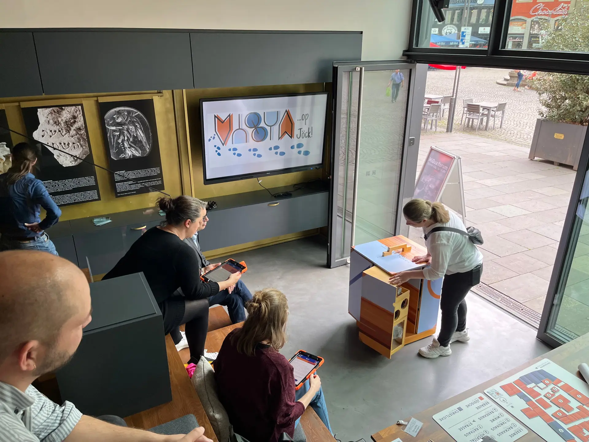 Eine Gruppe Menschen sitzt auf einer Treppe in einem Raum und schaut einem Vortrag zu.