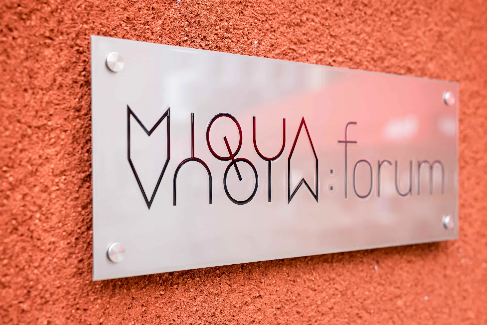 Ein silbernes Metallschild hängt an einer roten Außenwand.  Darauf steht MiQua:forum.
