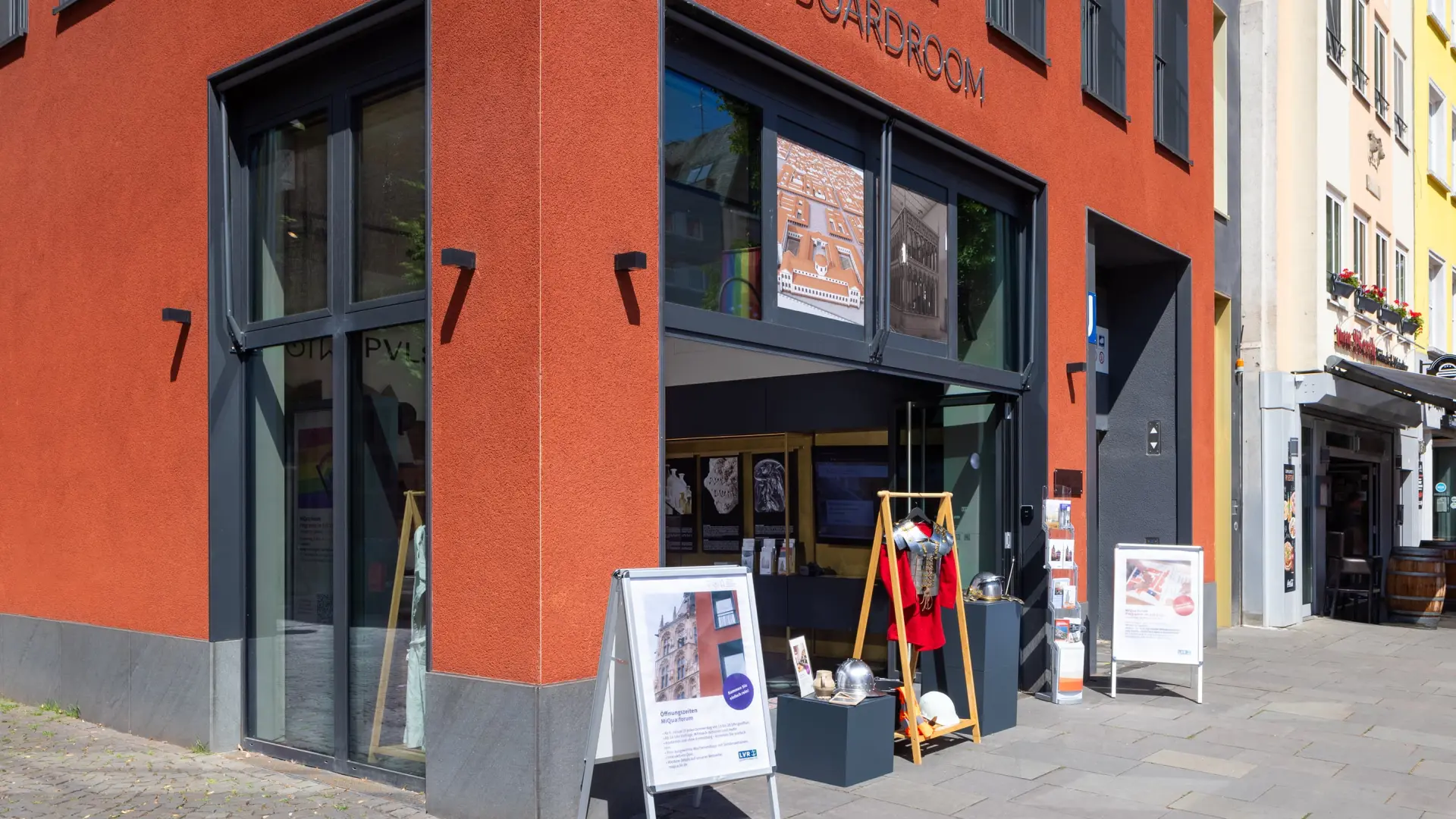 In einem roten Haus ist eine Fensterfront zu sehen, die geöffnet ist. Davor stehen zwei Kundenstopper und das Oberteil eines Ritterpanzers.