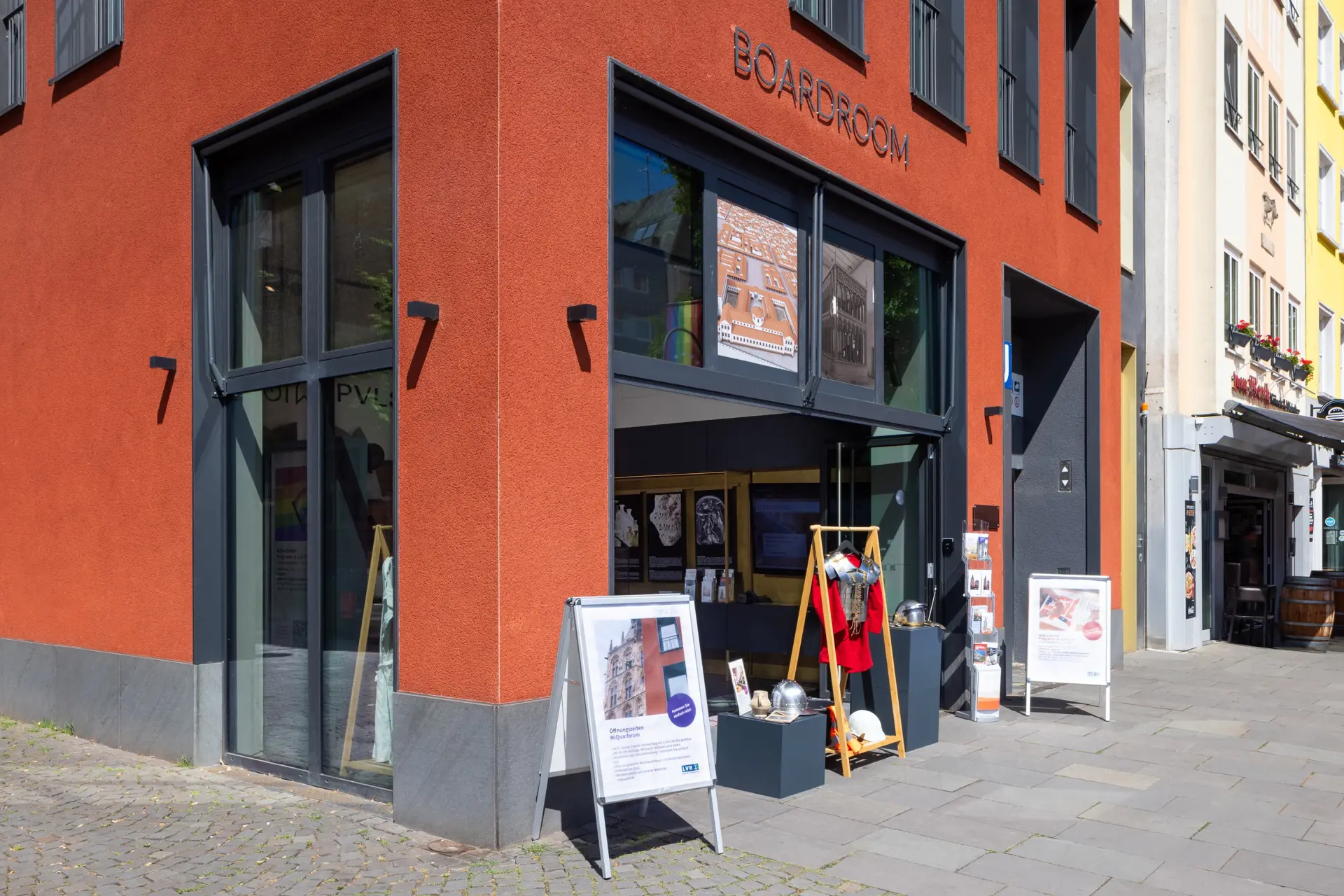 In einem roten Haus ist eine Fensterfront zu sehen, die geöffnet ist. Davor stehen zwei Kundenstopper und das Oberteil eines Ritterpanzers.