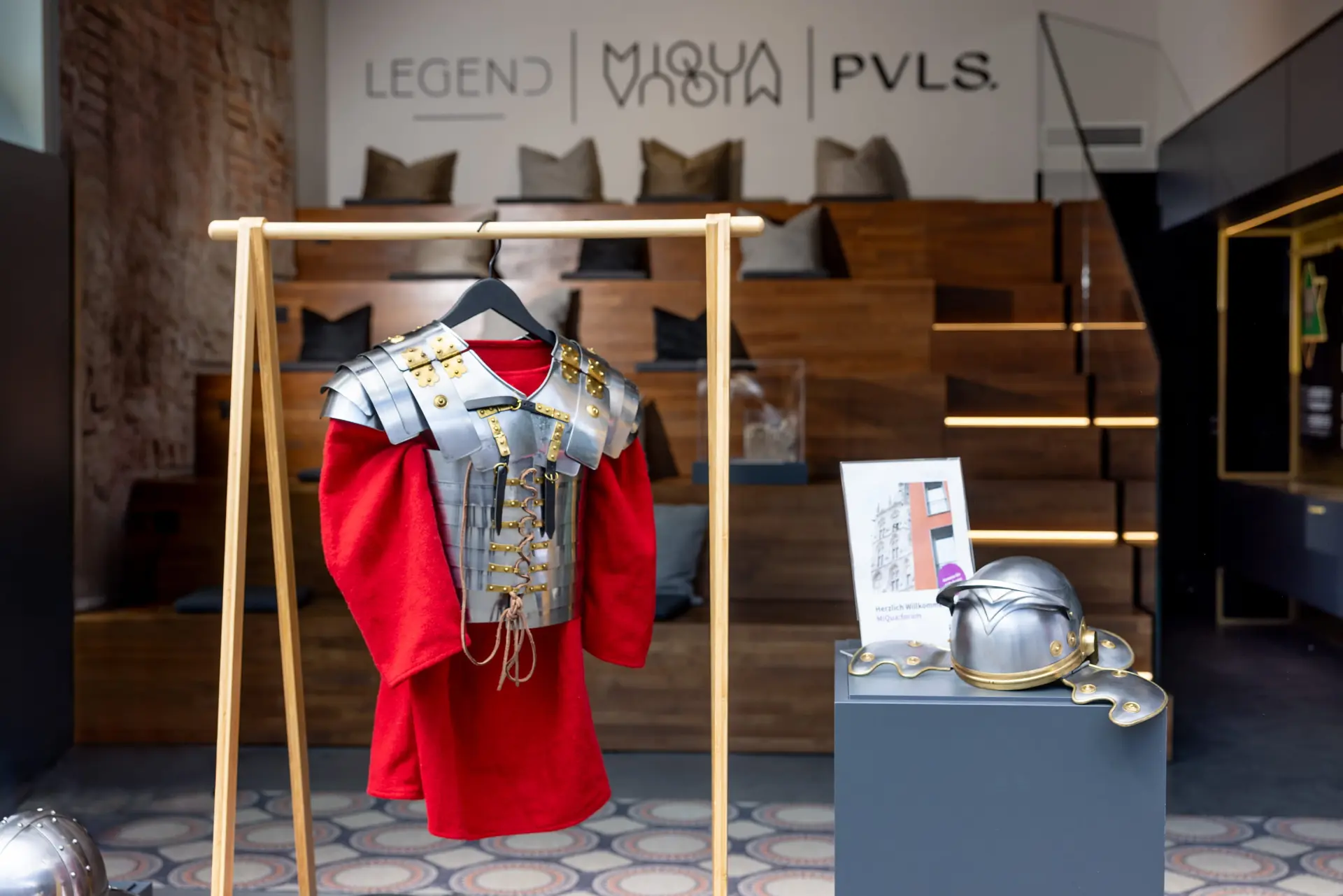 Das Oberteil einer Ritterrüstung hängt an einem Gestell.  Im Hintergrund ist ein Besucherraum zu sehen.