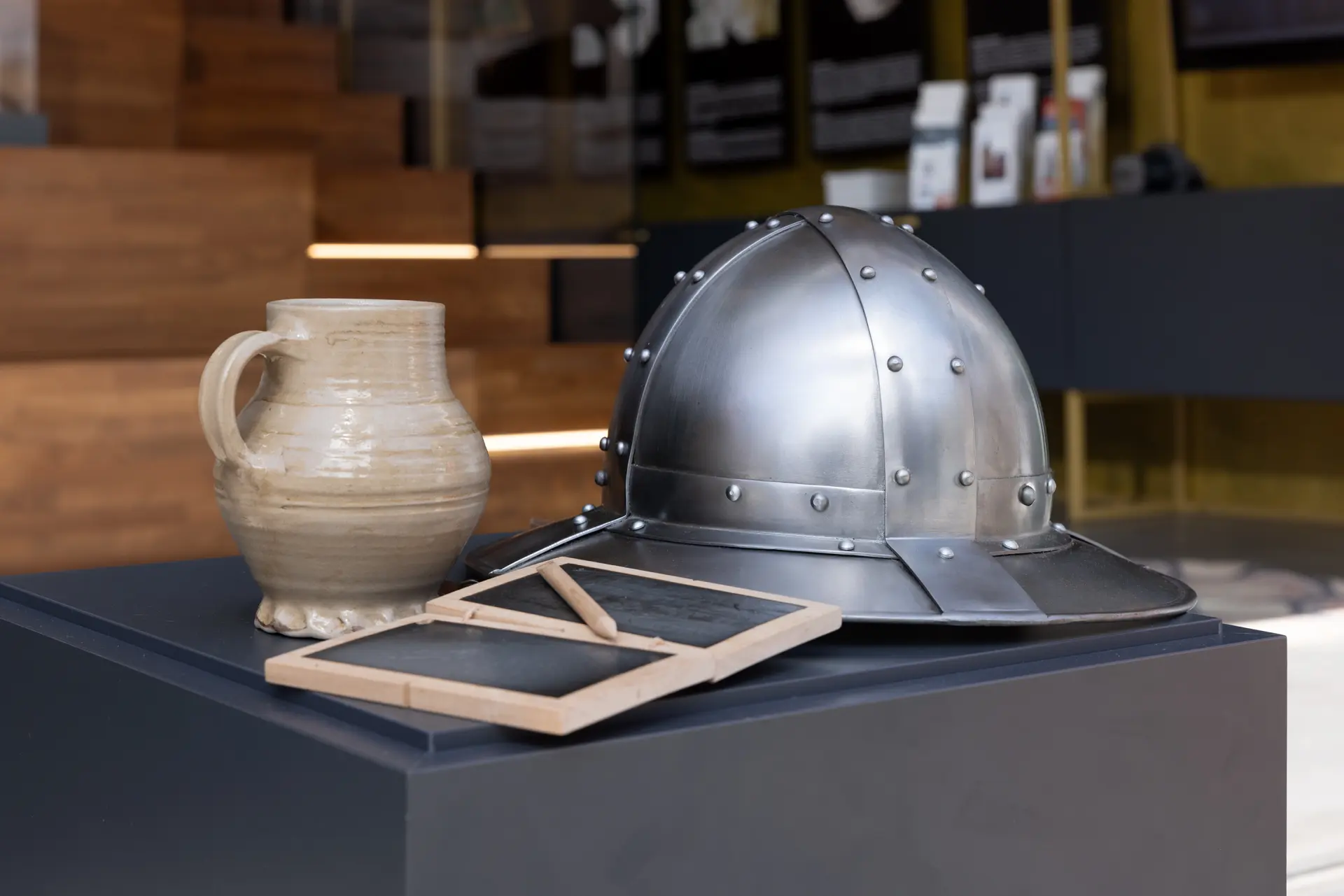 Ein Ritterhelm, ein Trinkkrug und eine Tafel zum schreiben stehen auf einem Podest.