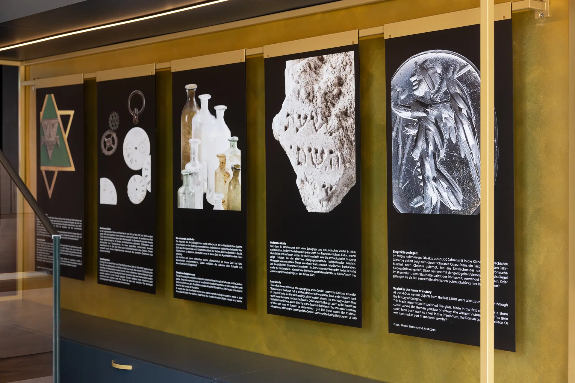 Fünf schwarze Informationstafeln sind nebeneinander aufgehängt.  Auf jeder Tafel ist ein Objekt aus den Ausgrabungen zu sehen.