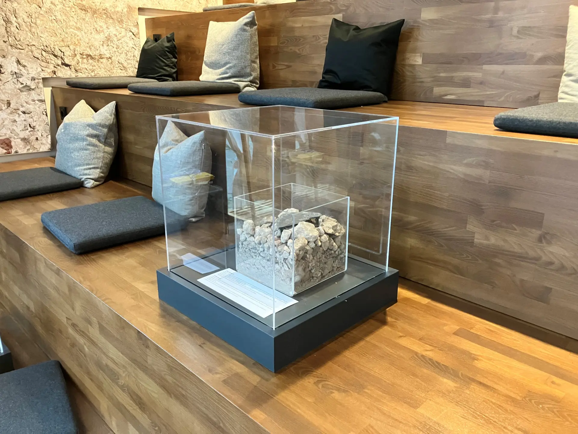 Eine Vitrine steht auf einer Treppe in einem Raum.  In der Vitrine ist ein Behälter aus Plexiglas, der Steine aus einer Ausgrabung beinhaltet.