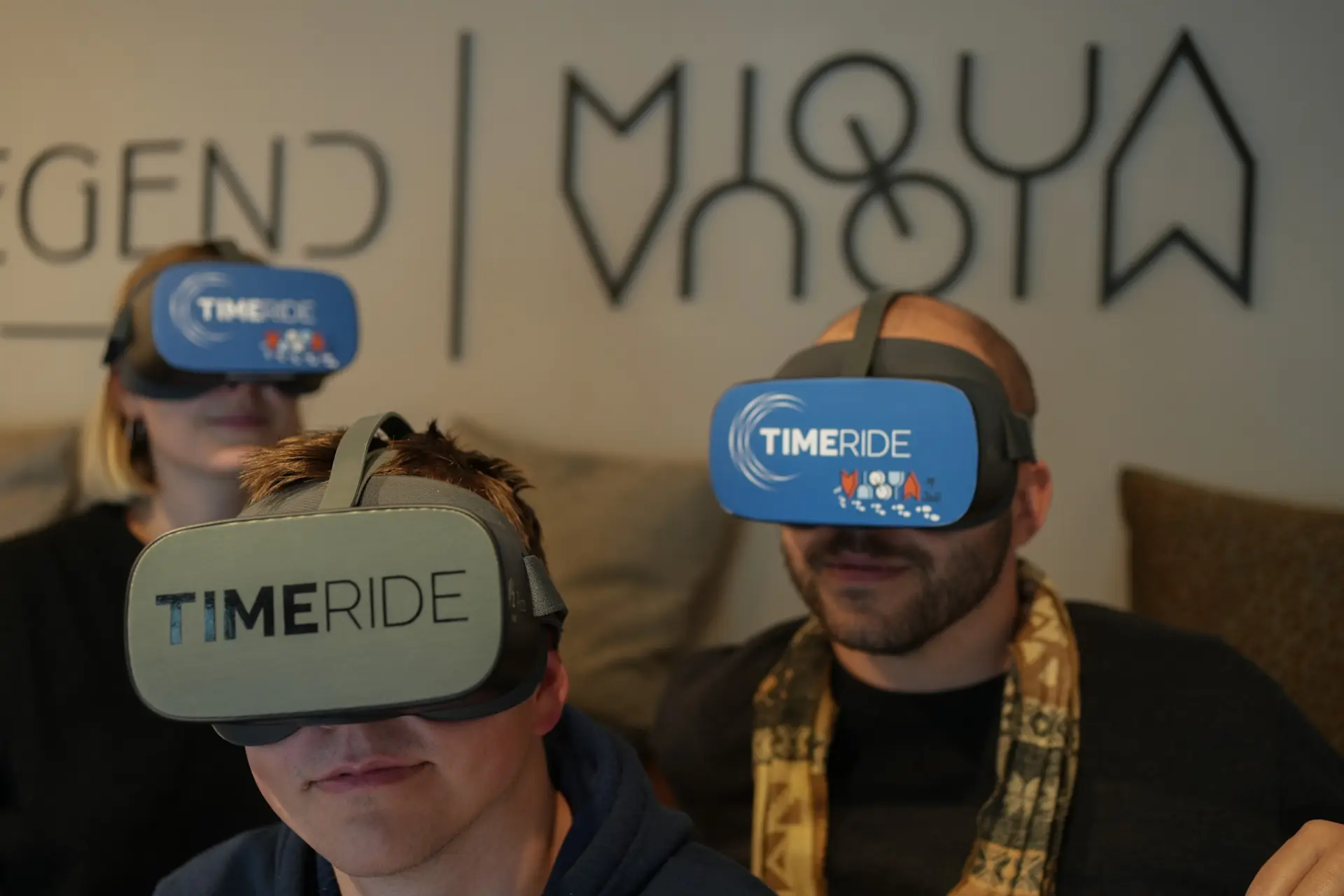 Drei Menschen sitzen in einem Raum und tragen dunkle Brillen, auf denen TimeRide drauf steht.