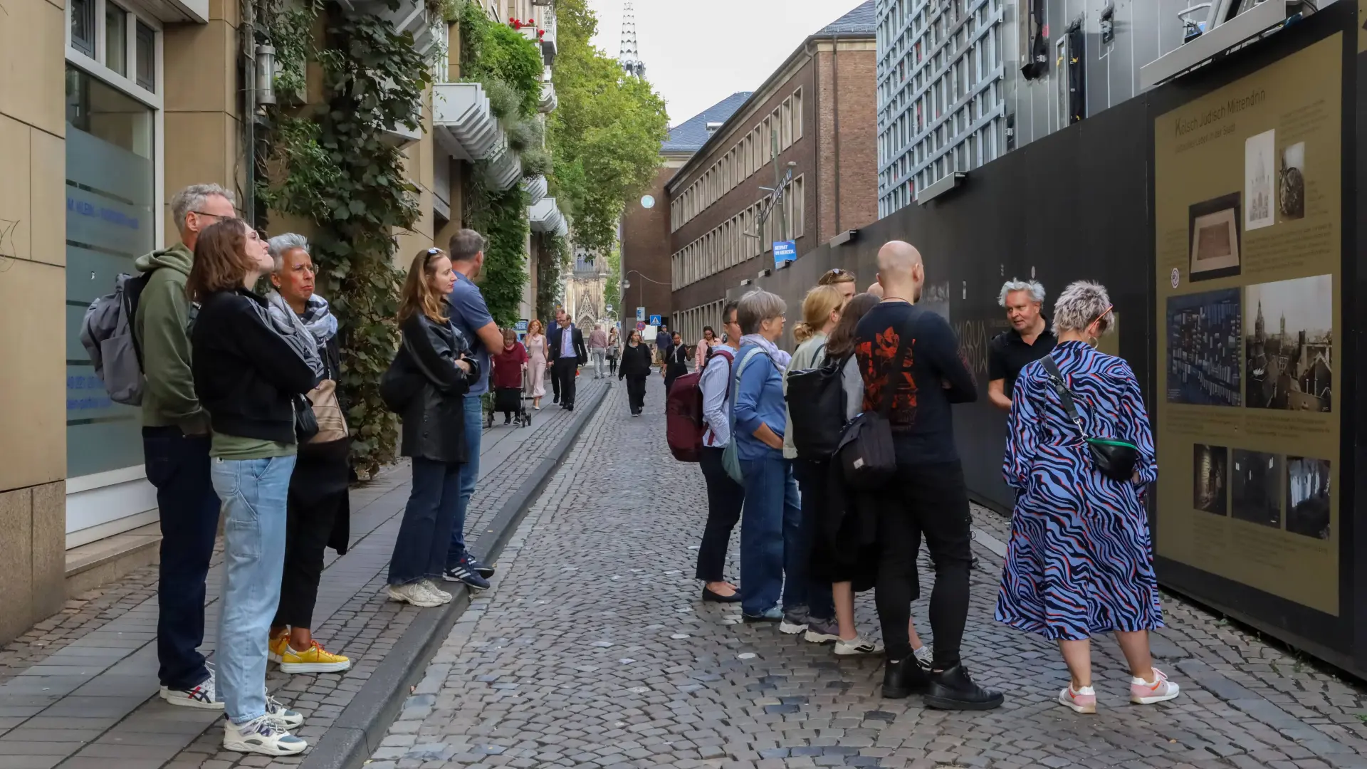 Eine Gruppe Menschen steht vor einem Bauzaun und hört einem Herren zu, der etwas erzählt.