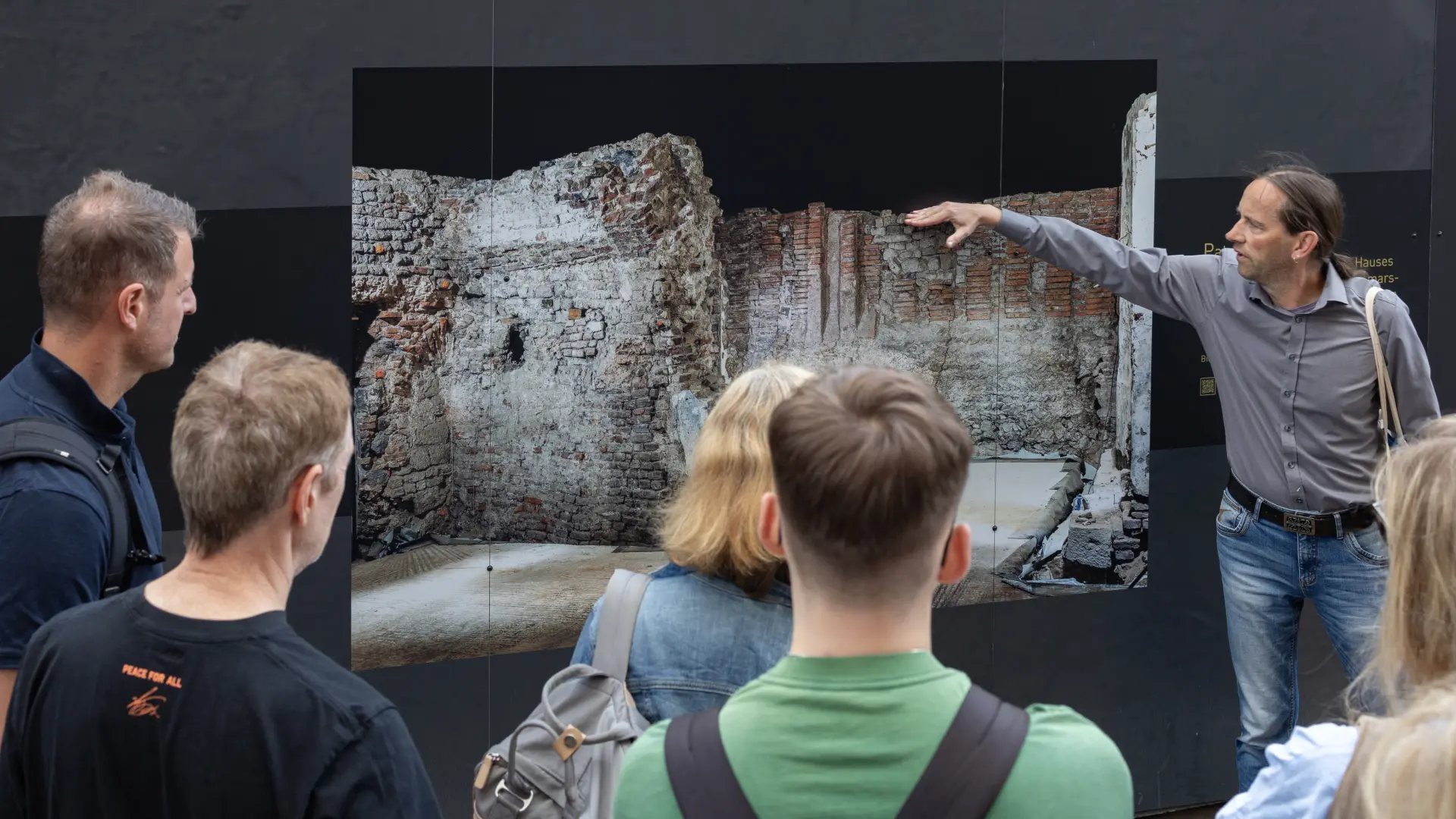 Ein Mann steht vor einem schwarzen Bauzaun und zeigt auf die Abbildung darauf.  Dort ist ein mittelalterlicher Kellerraum zu sehen.  Vor dem Bauzaun steht eine Gruppe Menschen, die dem Mann zuhören.