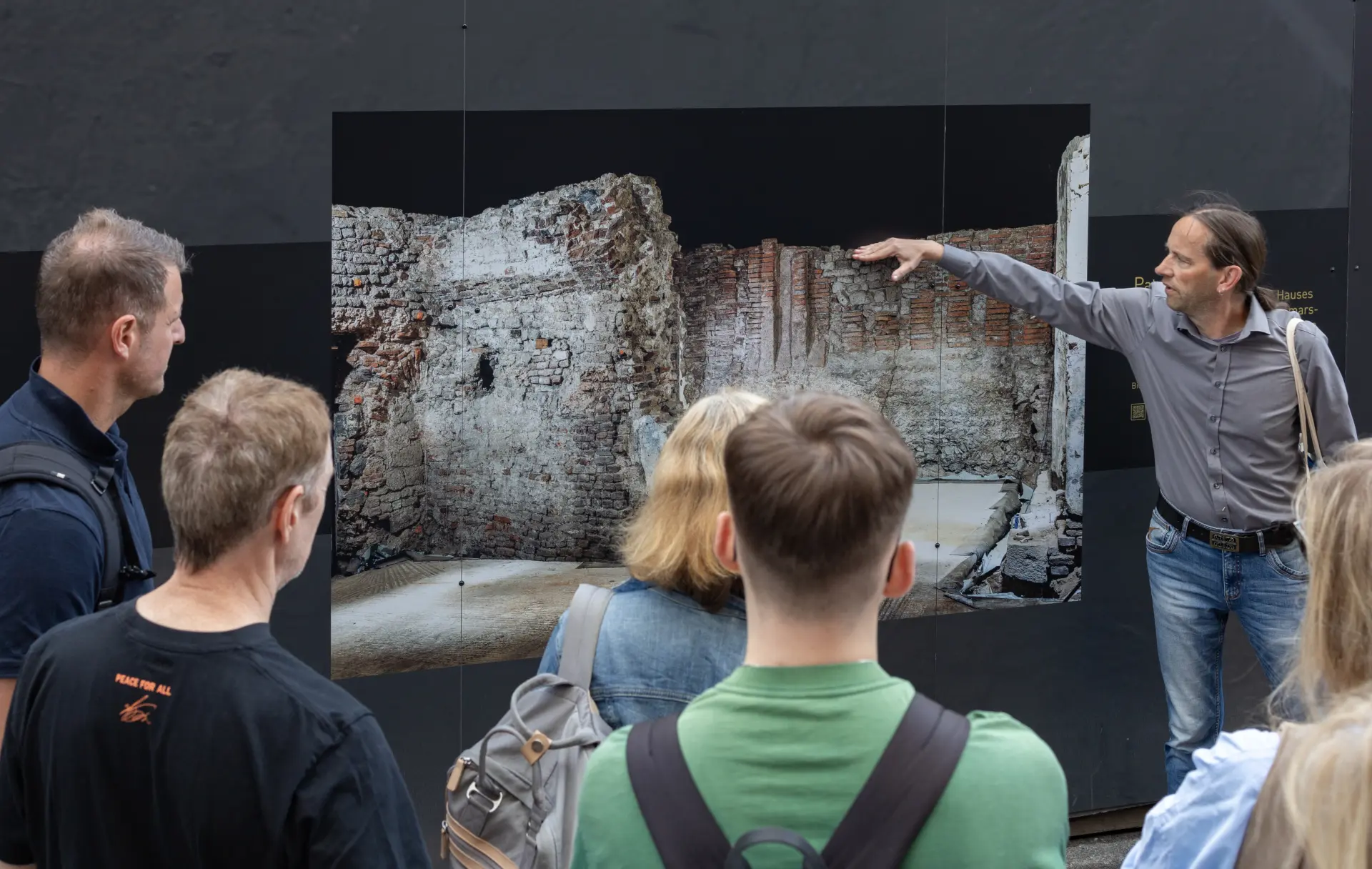 Ein Mann steht vor einem schwarzen Bauzaun und zeigt auf die Abbildung darauf.  Dort ist ein mittelalterlicher Kellerraum zu sehen.  Vor dem Bauzaun steht eine Gruppe Menschen, die dem Mann zuhören.