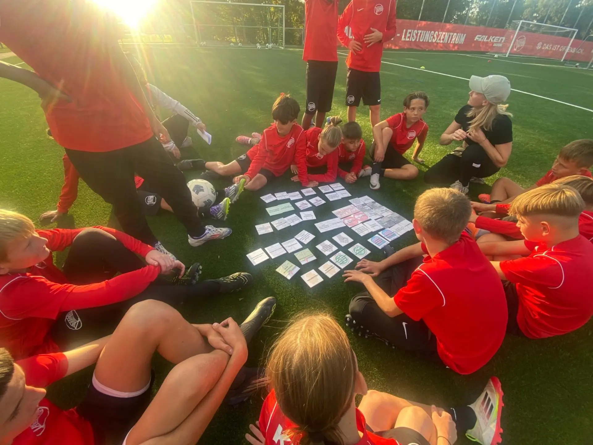 Eine Gruppe von Kindern in roten T-Shirts sitzen im Kreis auf einem Fußballfeld. In der Mitte liegen weiße Karten auf dem Boden.