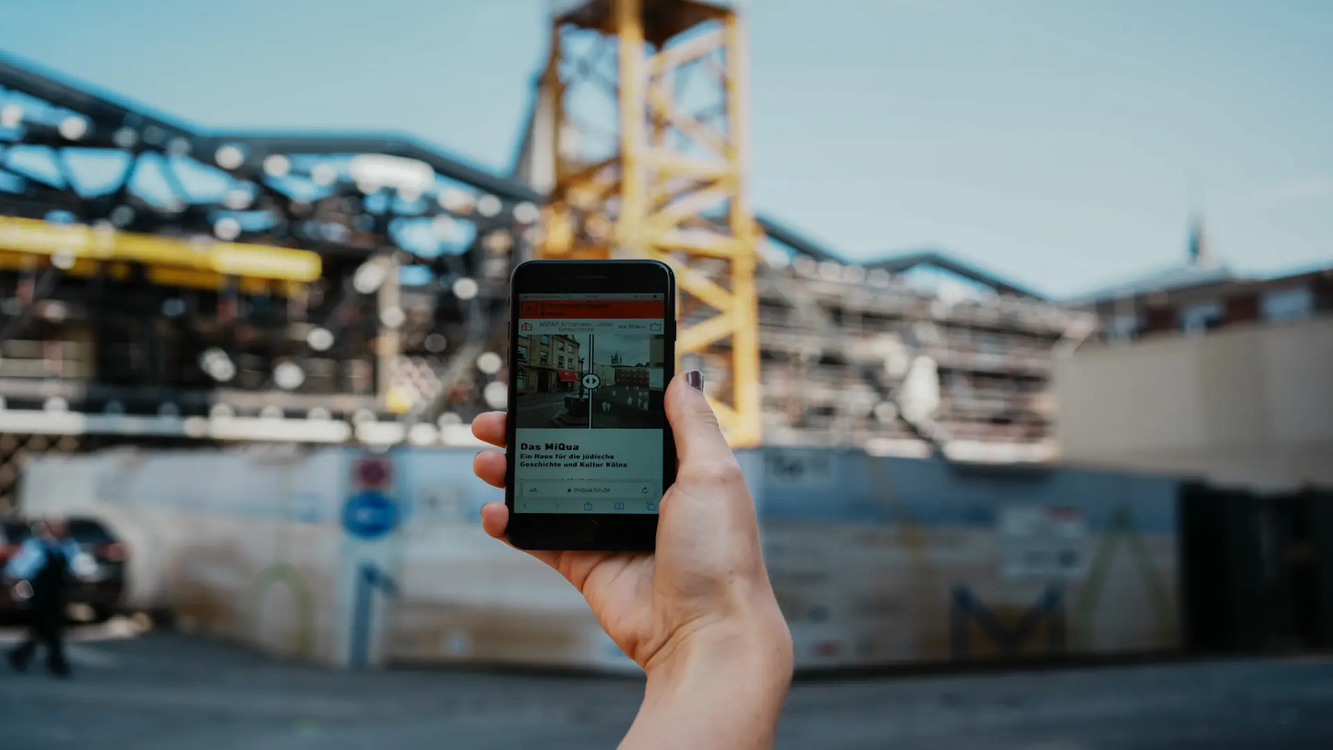 Eine Hand hält ein Handy hoch, auf dem man ein Foto und Text sehen kann.  Im Hintergrund ist eine Baustelle zu sehen.
