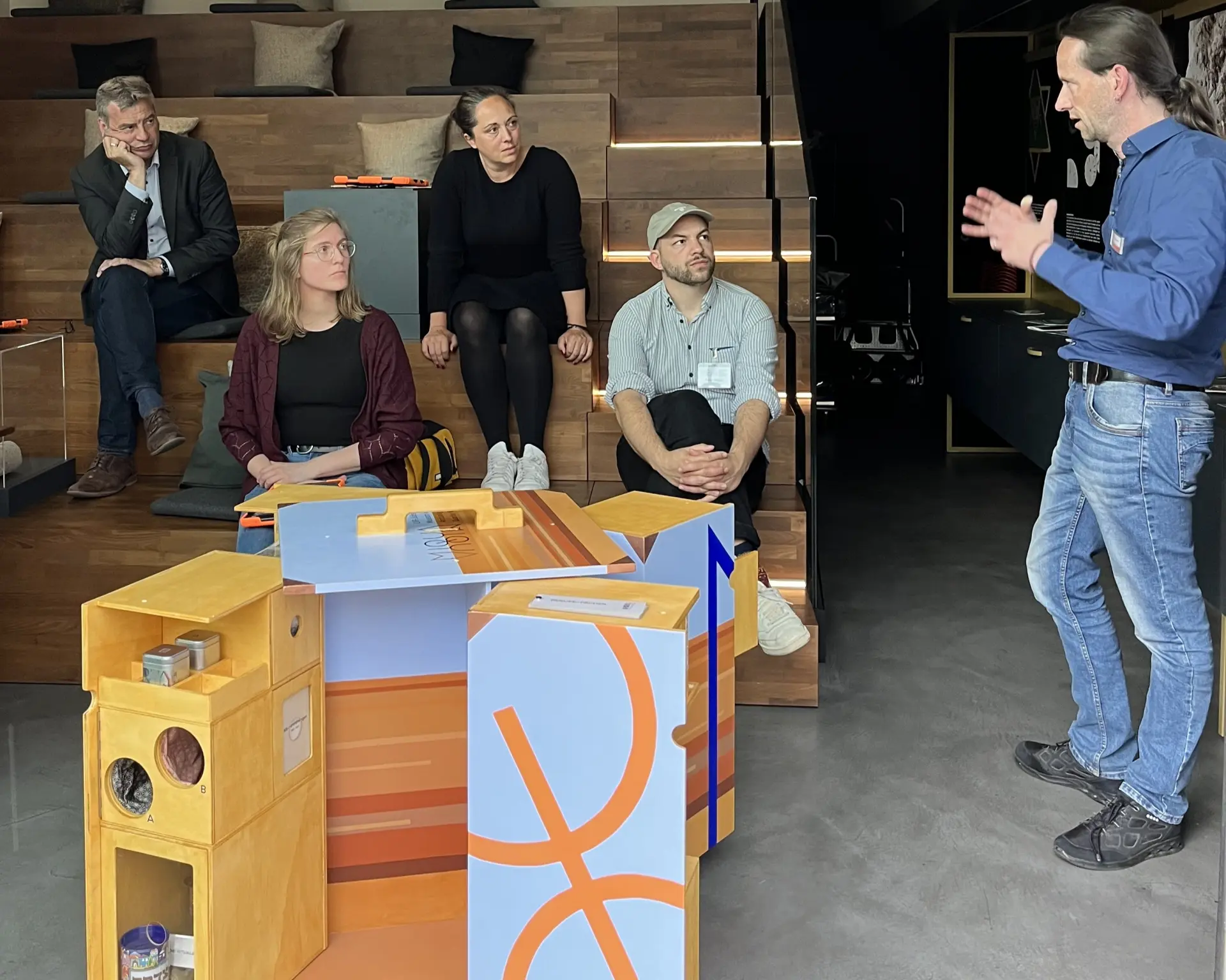 Ein Gruppe Menschen sitzt in einem Raum auf einer Treppe und schaut einem Herren zu, der ein hölzernes, aufgeklapptes Möbelstück in der Größe von einer Waschmaschine demonstriert.