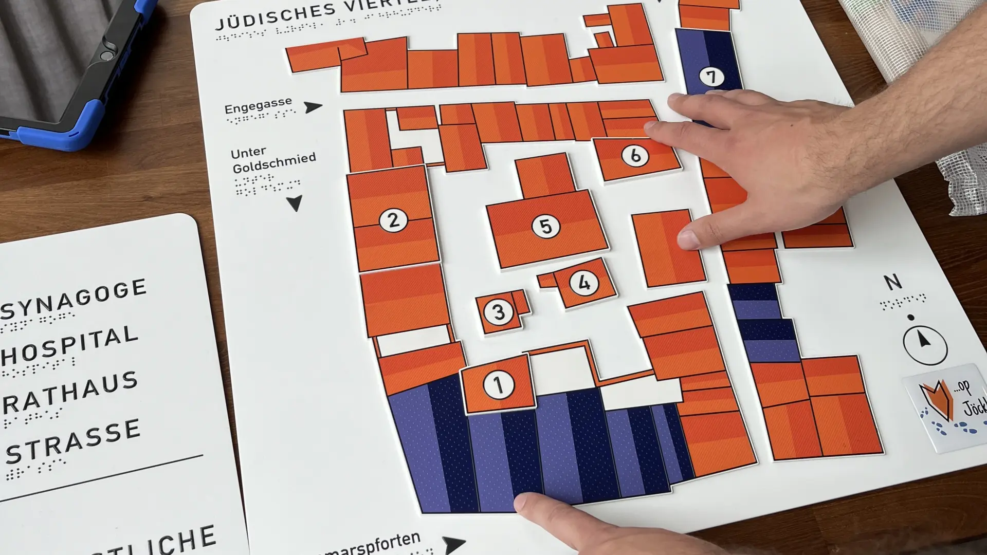 Ein Tastplan aus orangenen und lilafarbenen Rechtecken die angeordnet und nummeriert sind.  Auf einer Legende stehen Wörter wie Synagoge, Hospital und Rathaus.