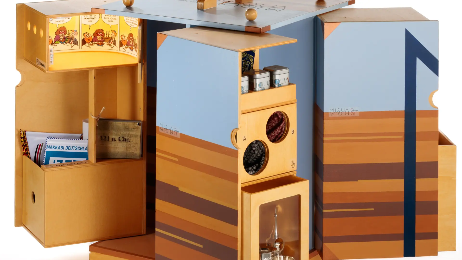 Ein Möbelstück aus holz ist in hellblau und orange bemalt, und so groß wie eine Waschmaschine.  Man kann es an den Seiten öffnen und auseinanderziehen.  Darauf steht ein Tablet, wo „MiQua" drauf steht.