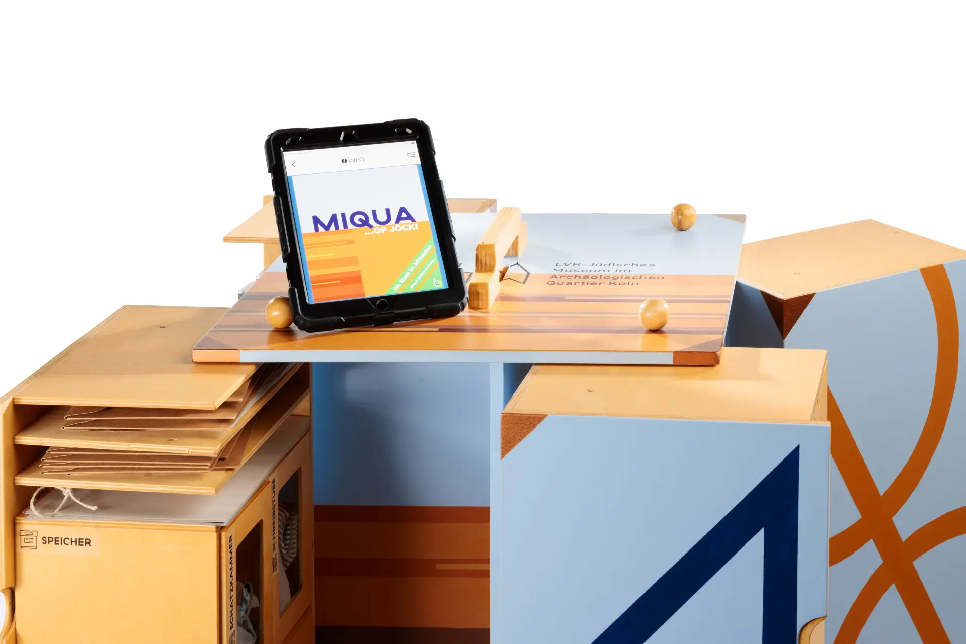 Ein Möbelstück aus holz ist in hellblau und orange bemalt, und so groß wie eine Waschmaschine.  Man kann es an den Seiten öffnen und auseinanderziehen.  Darauf steht ein Tablet, wo „MiQua" drauf steht.