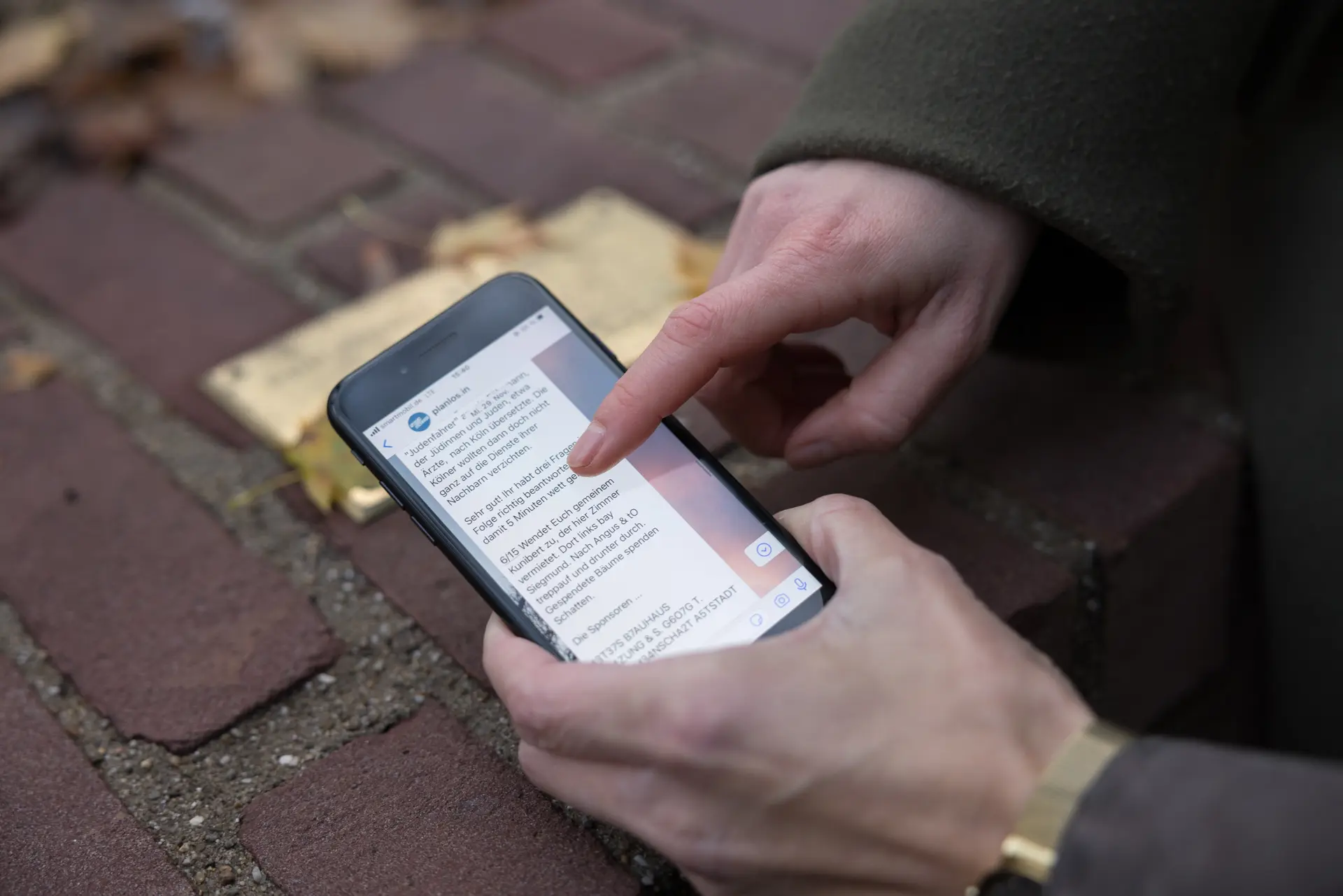 Eine Hand hält ein Handy.  Im Hintergrund sieht man eine Steinmauer, auf der eine goldene Plakette befestigt ist.