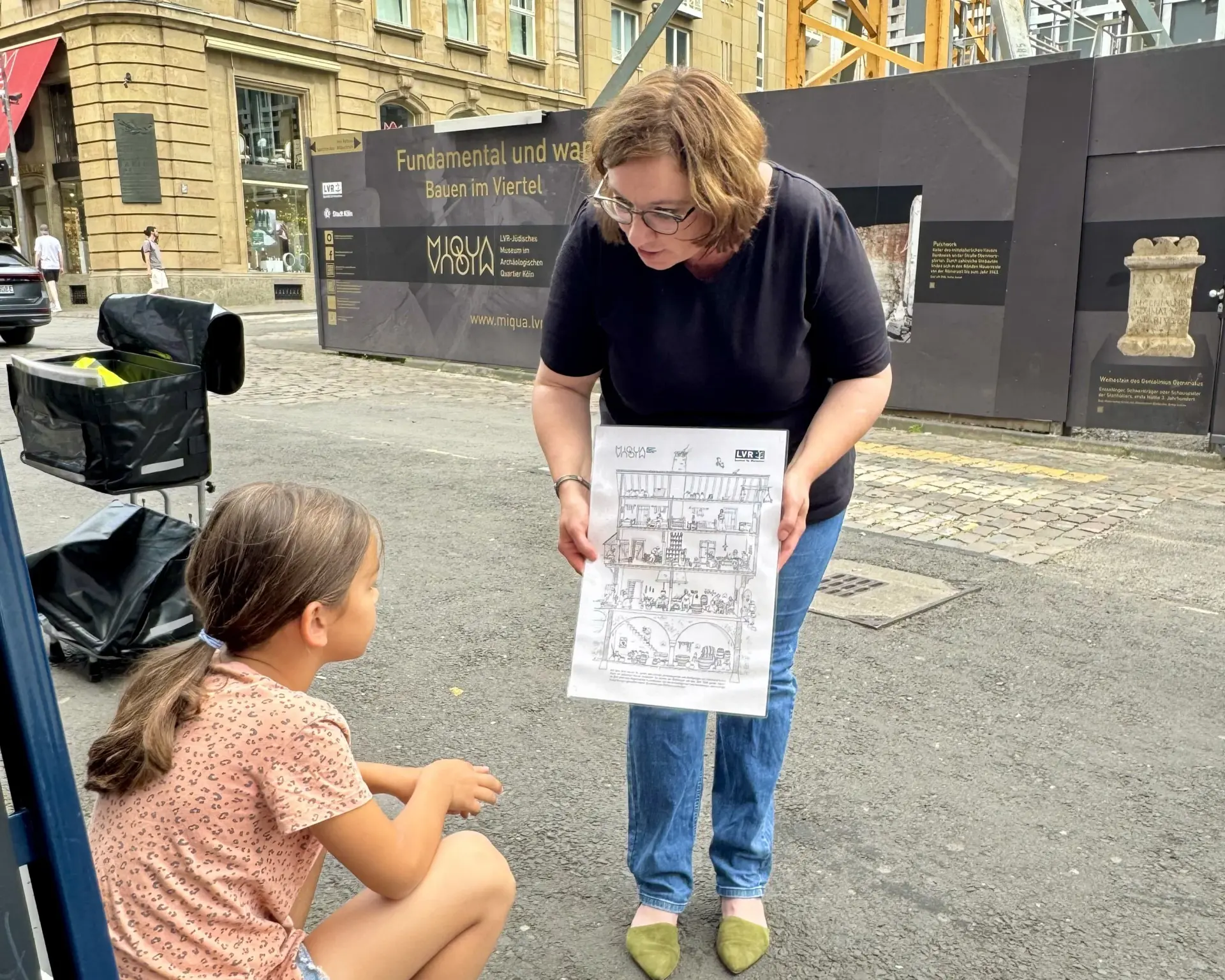 Eine erwachsene Frau hält eine Grafik, die von einem Mädchen bestaunt wird.  Im Hintergrund ist eine Baustelle zu sehen.