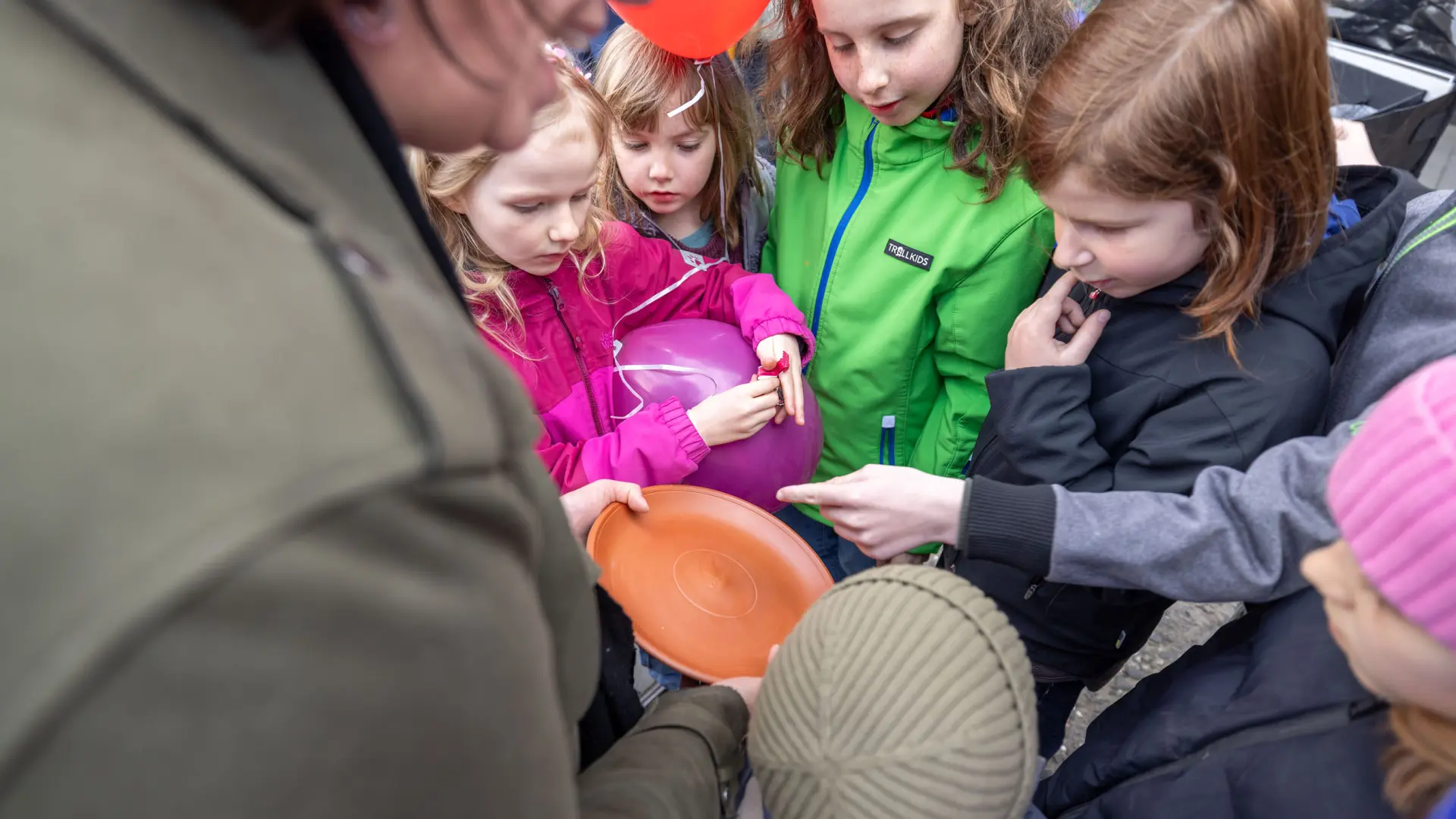 Eine Gruppe Kinder steht im Kreis und zeigt auf ein Objekt, dass eine Erwachsene hält.