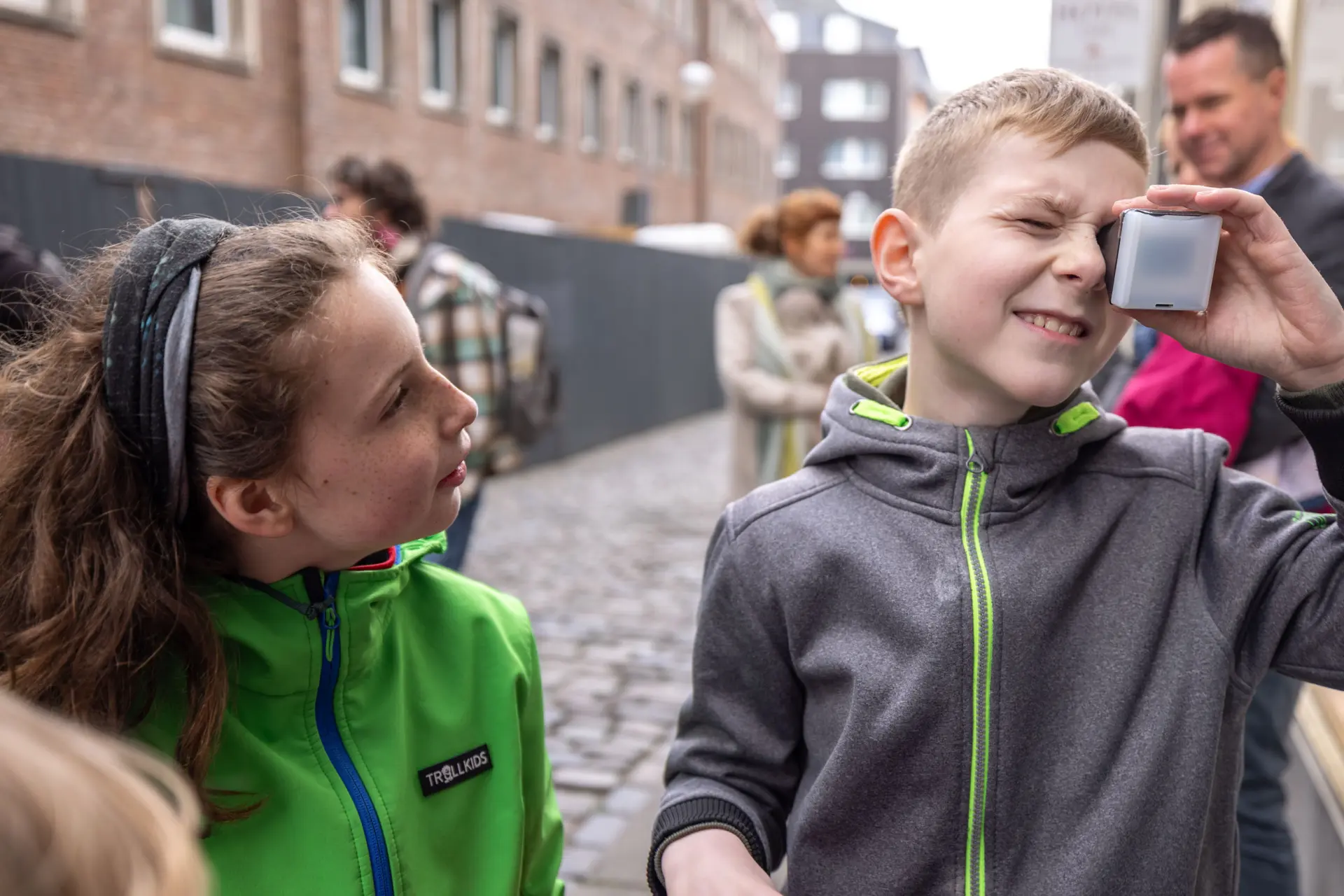 Ein Junge schaut durch ein kleines Kästchen.  Neben ihm steht ein Mädchen, das ihn ansieht.