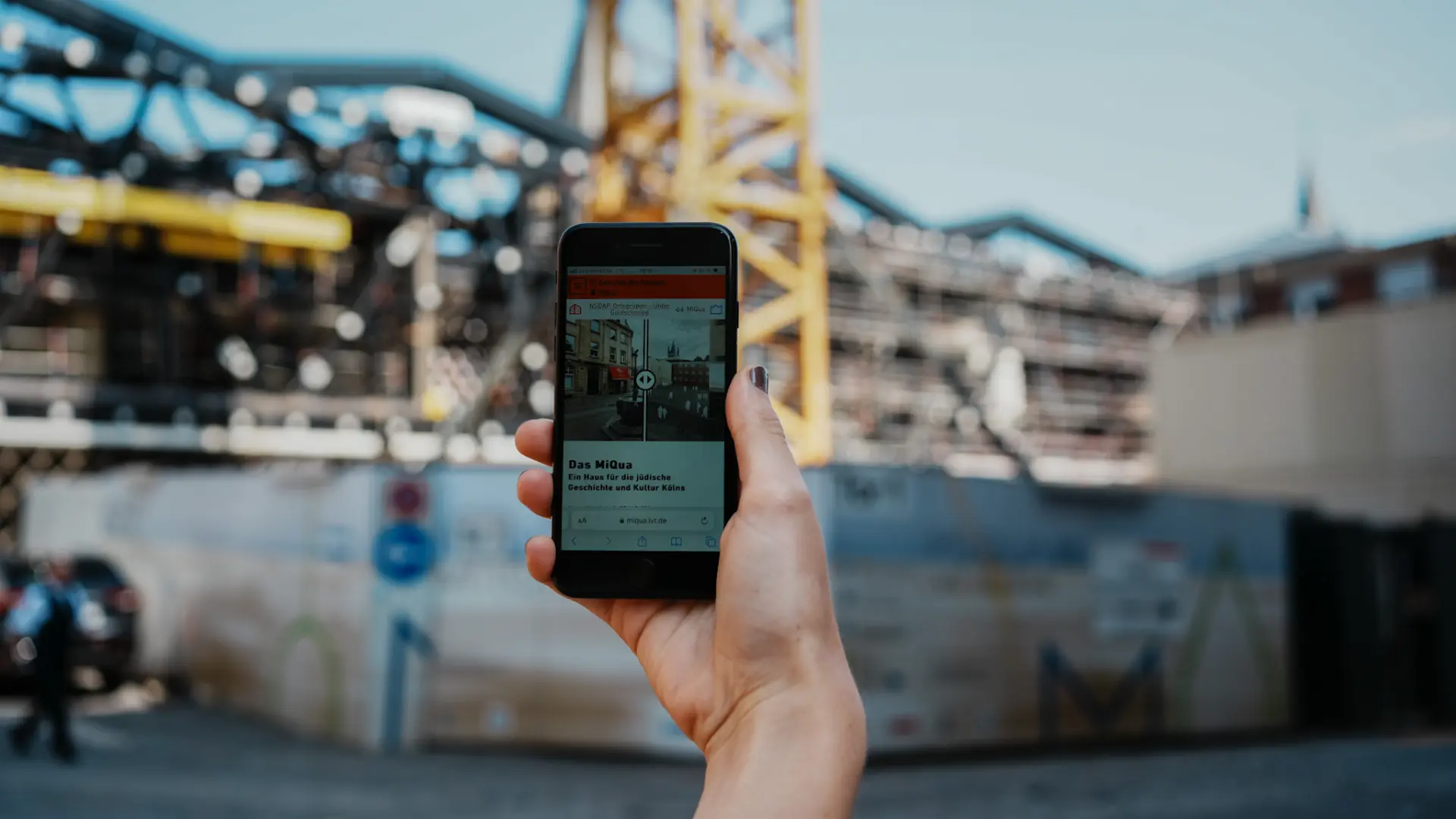 Eine Hand hält ein Handy in die Luft, auf der eine App zu sehen ist. Im Hintergrund ist eine Baustelle zu sehen.