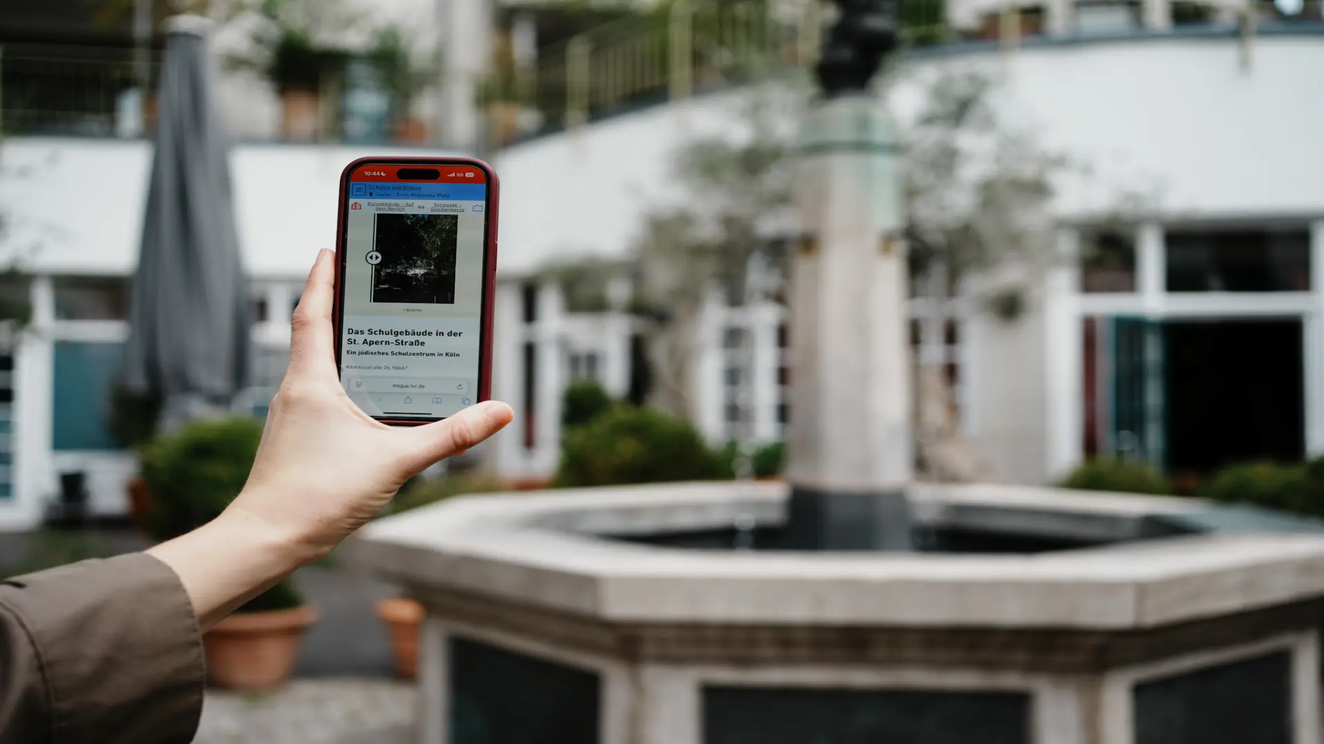 Eine Hand hält ein Handy in die Luft.  Im Hintergrund ist ein Springbrunnen zu sehen.