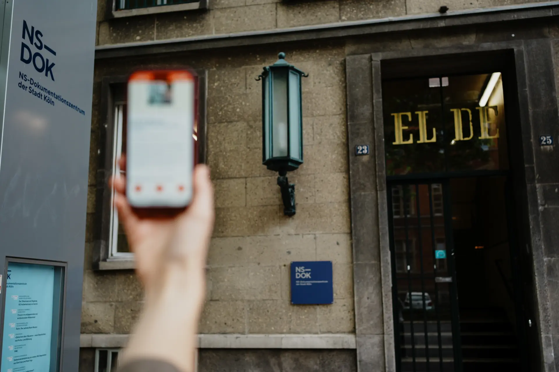 Eine Hand hält ein Handy hoch auf der eine App zu sehen ist.  Im Hintergrund ist ein Haus zu sehen mit der Aufschrift El-De.