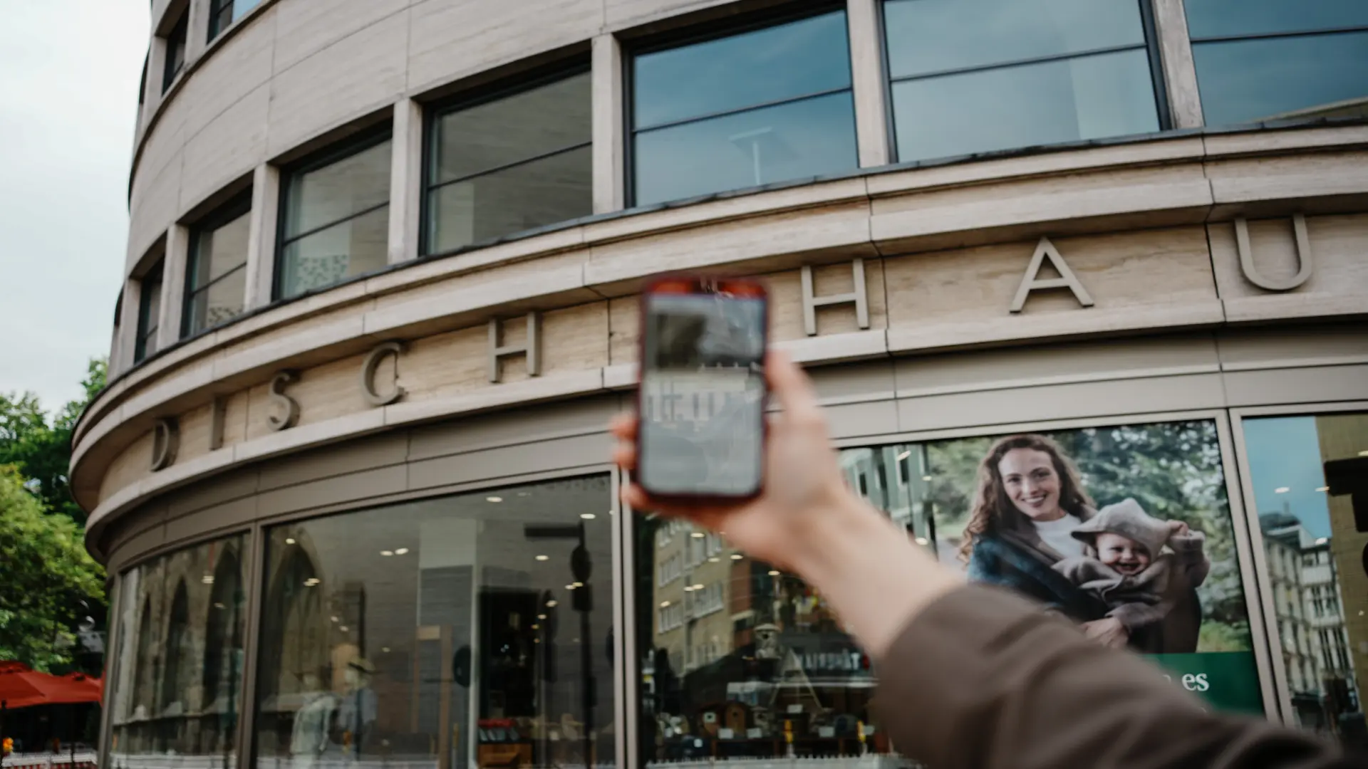 Eine Hand hält ein Handy in die Luft, auf dem eine App zu sehen ist.  Im Hintergrund sieht man ein Haus mit der Aufschrift Disch Haus.