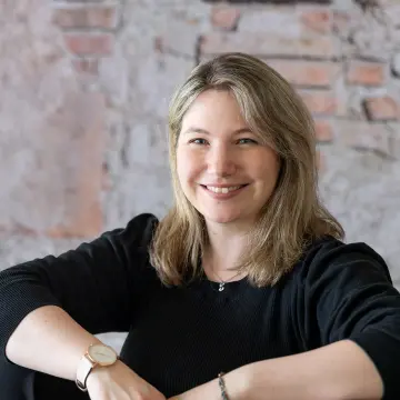 Eine Mitarbeiterin von MiQua sitzt auf einer Holztreppe.  Sie trägt schwarze Kleidung und lächelt in die Kamera.