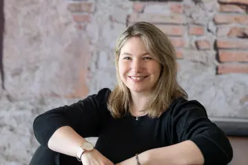 Bild: Eine Mitarbeiterin von MiQua sitzt auf einer Holztreppe. Sie trägt schwarze Kleidung und lächelt in die Kamera.