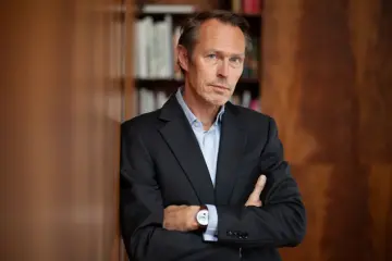 Ein Herr im Anzug hat die Arme übereinandergelegt und lehnt gegen eine Wand die mit Holz verkleidet ist.