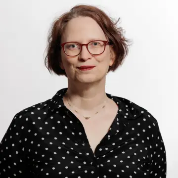 Portrait-Foto einer Mitarbeiterin von MiQua vor dunklem Hintergrund. Sie trägt ein weißes Hemd, darüber einen dunklen Blazer und eine Brille. Ihre rötlichen Haare hat sie zu einem Zopf gebunden. 