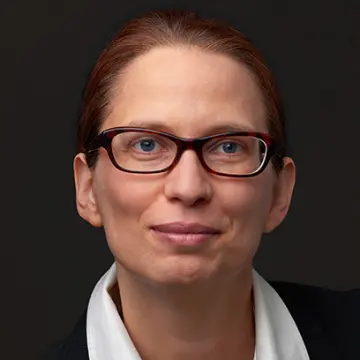 Portrait-Foto einer Mitarbeiterin von MiQua vor dunklem Hintergrund. Sie trägt ein weißes Hemd, darüber einen dunklen Blazer und eine Brille. Ihre rötlichen Haare hat sie zu einem Zopf gebunden. 