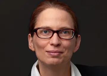Portrait-Foto einer Mitarbeiterin von MiQua vor dunklem Hintergrund. Sie trägt ein weißes Hemd, darüber einen dunklen Blazer und eine Brille. Ihre rötlichen Haare hat sie zu einem Zopf gebunden. 