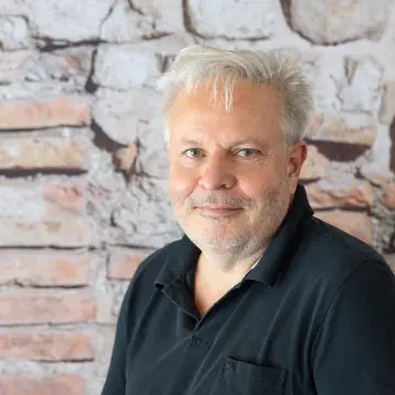 Ein Mann mit weißem Haar und kurzem Bart sitzt vor einer Backsteinfassade.