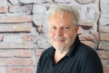 Ein Mann mit weißem Haar und kurzem Bart sitzt vor einer Backsteinfassade.