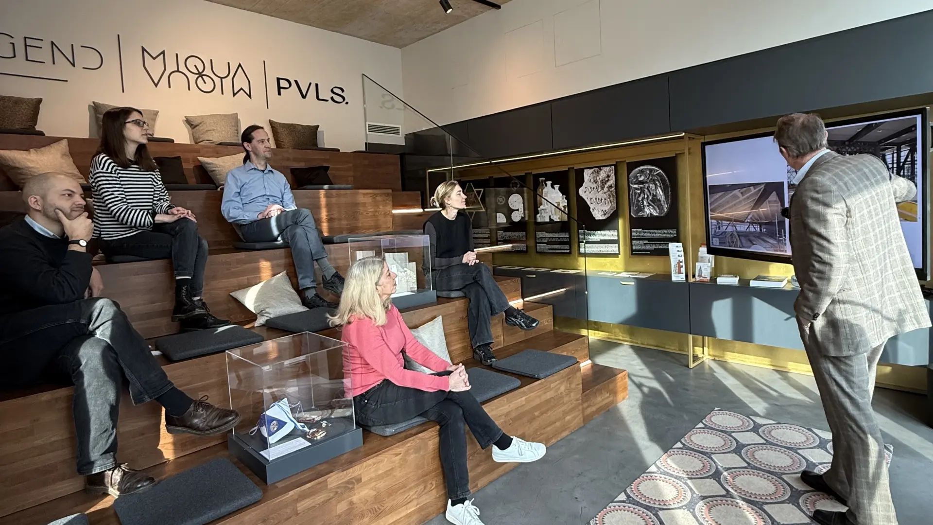 Eine Gruppe Menschen sitzen in einem Raum auf einer Treppe und hören einem Herren zu, der einen Vortrag hält und auf einen Bildschirm zeigt.