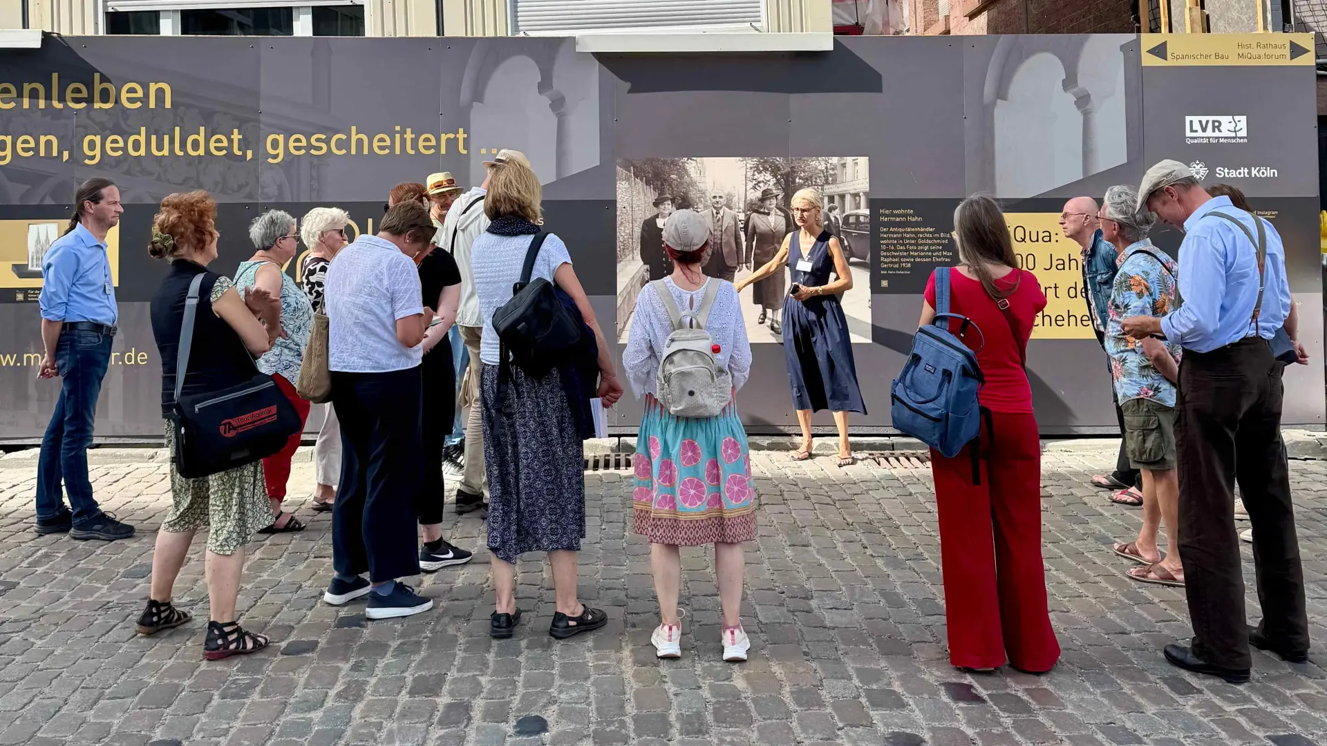 Eine Gruppe Menschen nimmt an einer Führung teil und steht vor einem Bauzaun.
