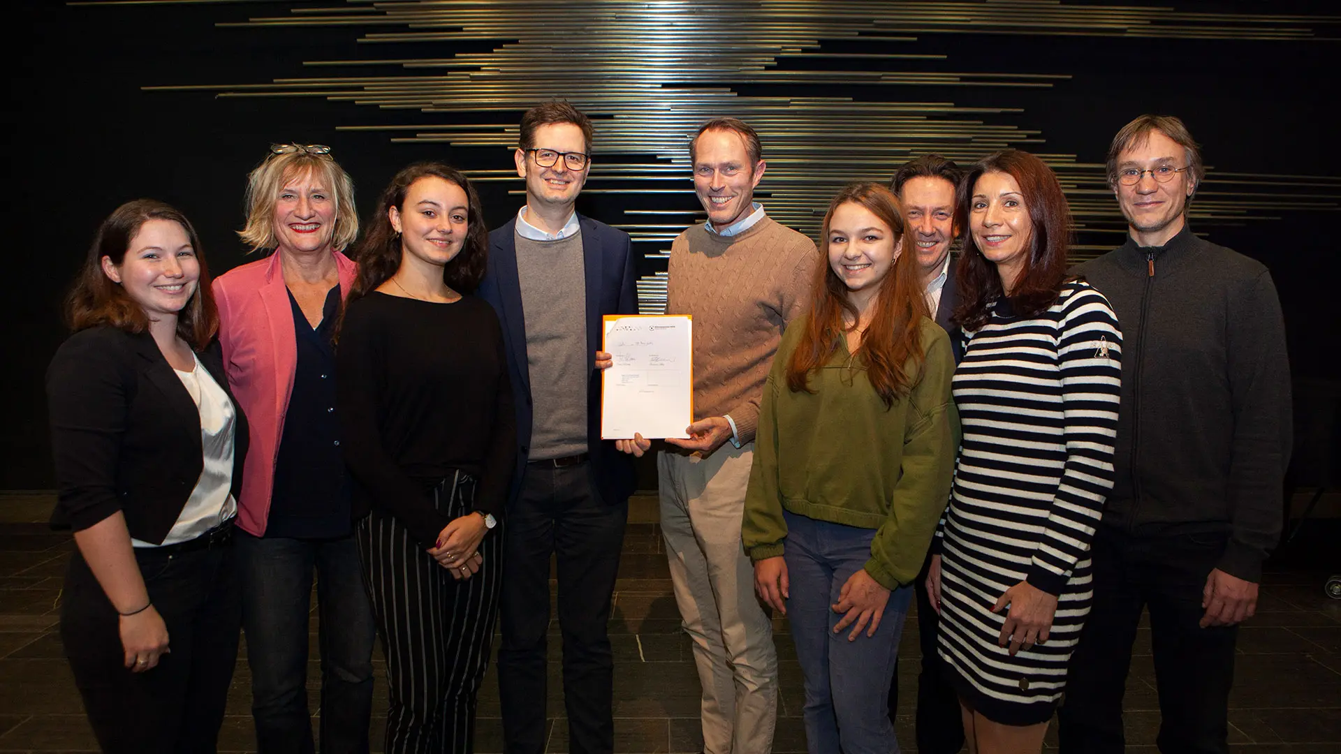 Ein Teil des MiQua-Teams sowie des Apostelgymnasiums Köln stehen im Halbkreis und zeigen stolz die unterschriebene Bildungspartnerschaft.