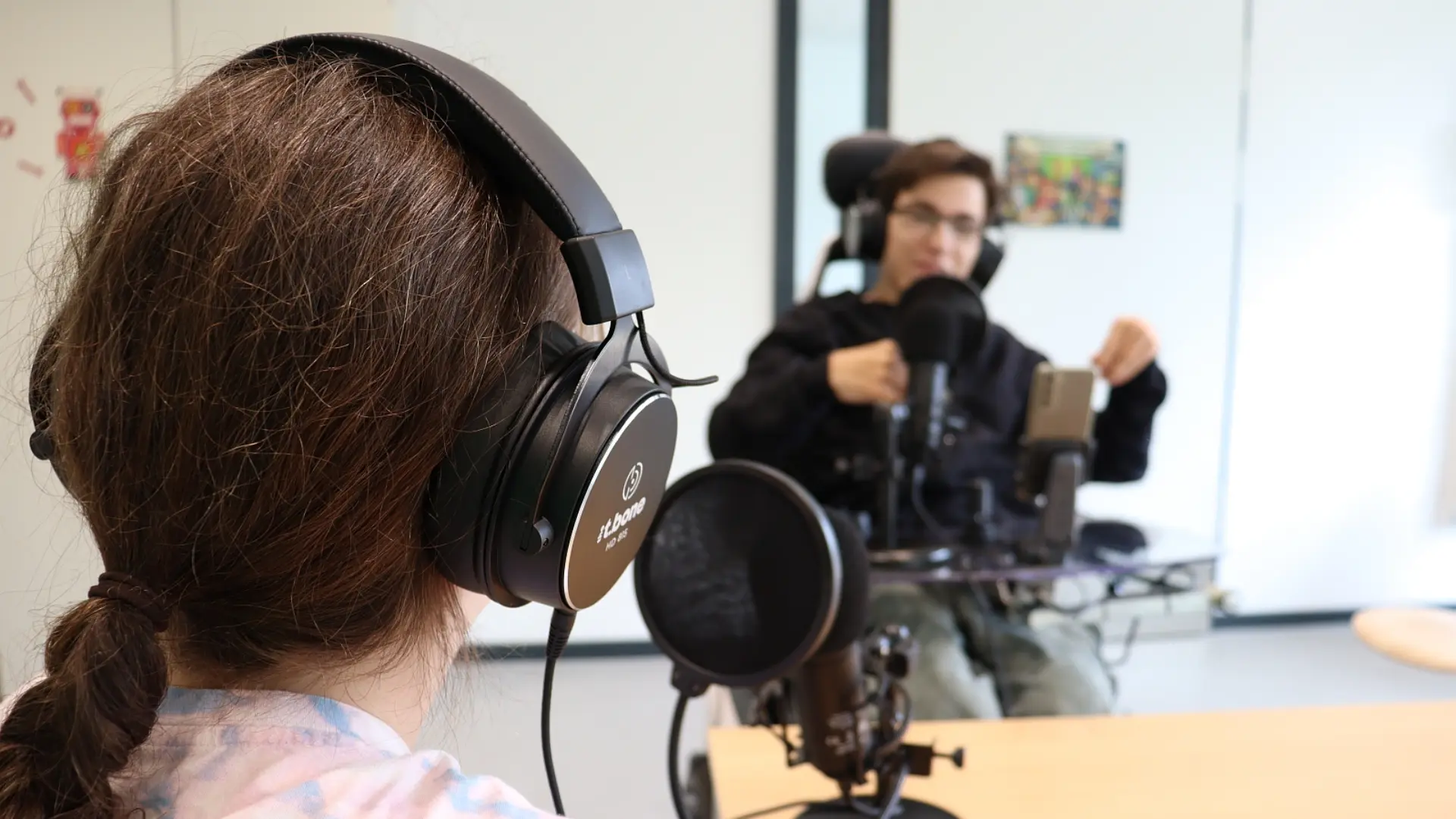 Zwei Jugendliche (eine im Rollstuhl) nehmen einen Podcast auf. Sie tragen große Kopfhörer und haben Mikrophone vor sich.
