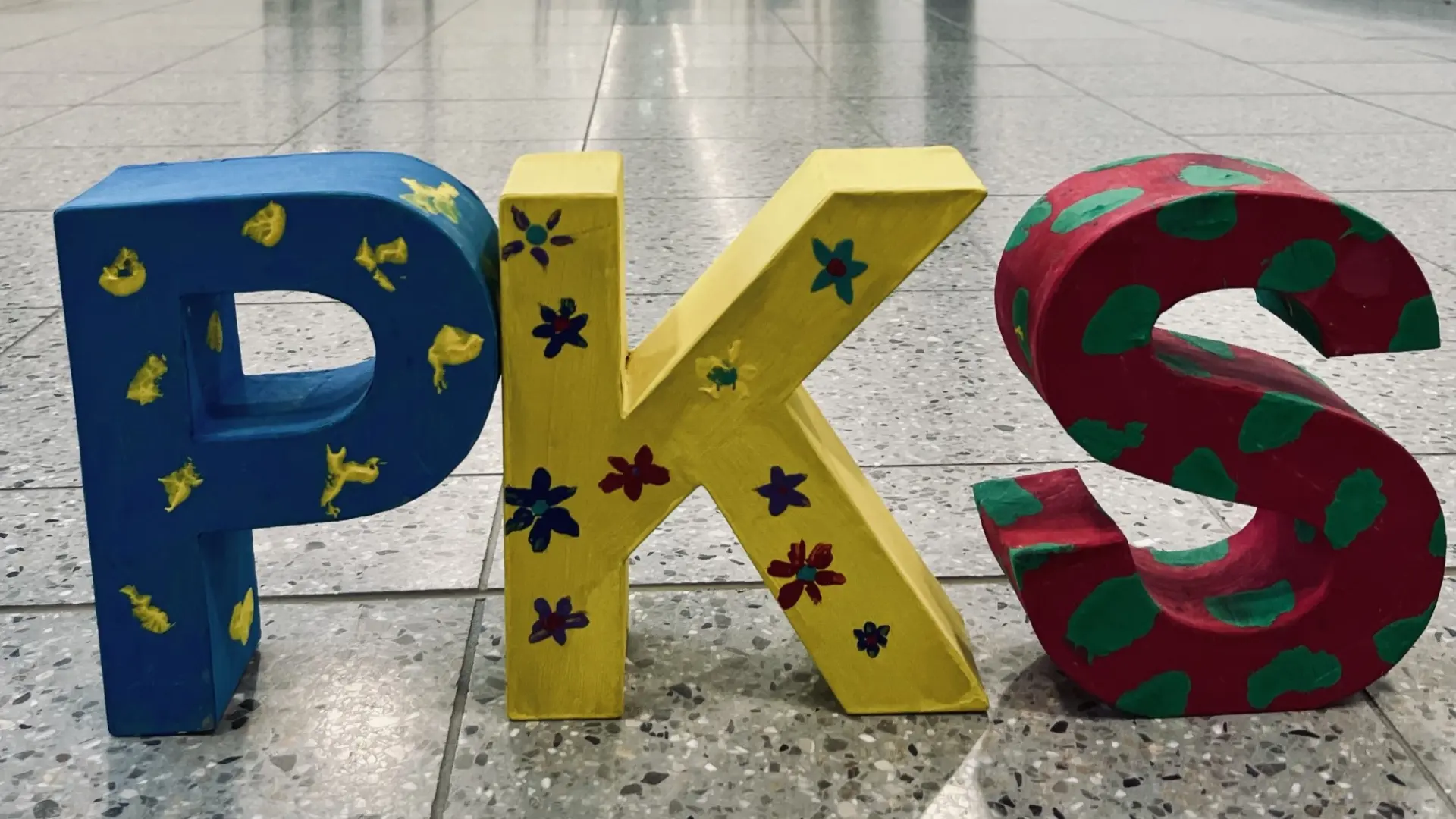 Die drei BuchstabenP, K und S aus bunt bemalter Pappe stehen auf dem Fußboden eines langen, hellen Flurs mit vielen abgehenden Türen.
