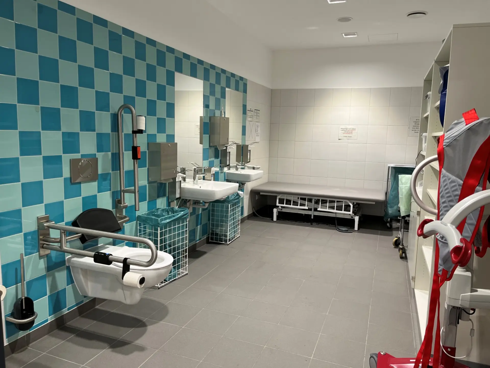 Ein blau gekachelter Sanitärraum. An der Wand stehen eine Liege und ein Toilettenlift.