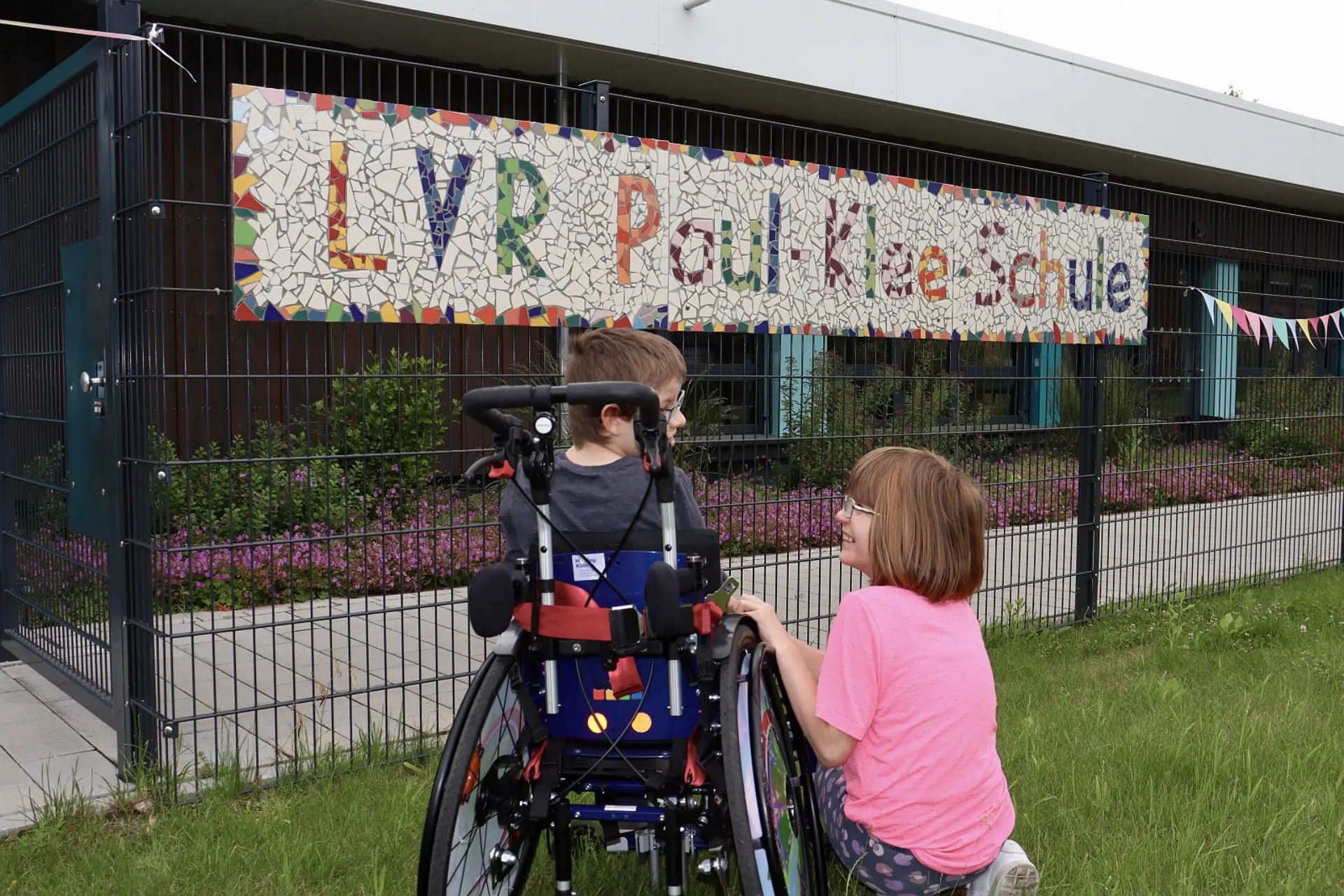 An einem Gitterstabzaun vor einem blauen Gebäude hängt ein Schild auf dem der aus bunten Mosaiksteinchen geformte Name "LVR-Paul-Klee-Schule" zu sehen ist.
