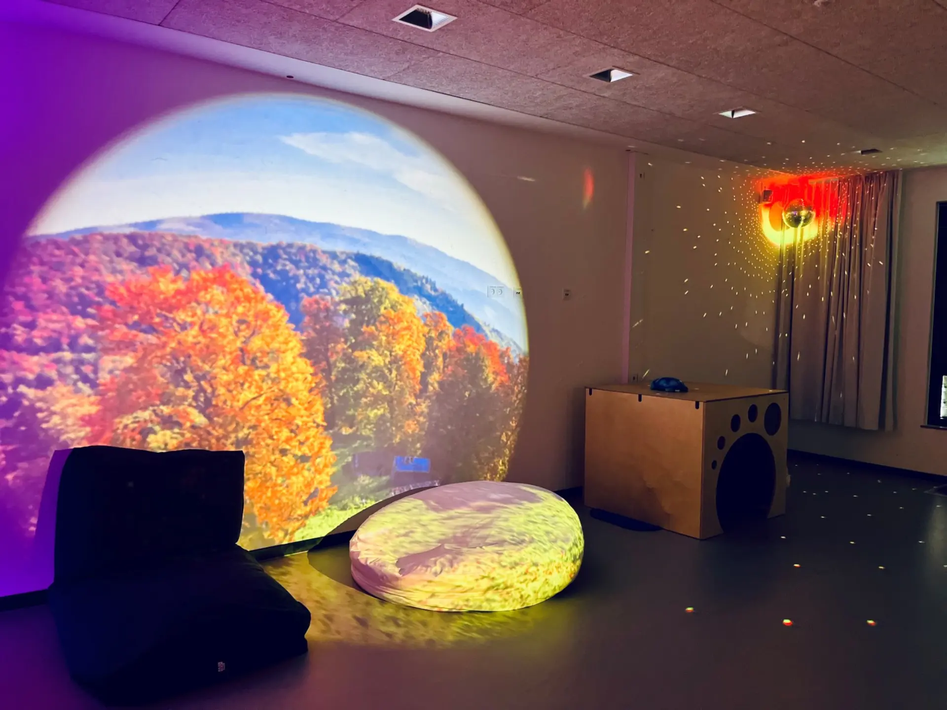 Snoezelen-Raum mit großer, runder Projektion einer herbstlichen Berglandschaft, Sitzkissen und einer glitzernden Spiegelkugel