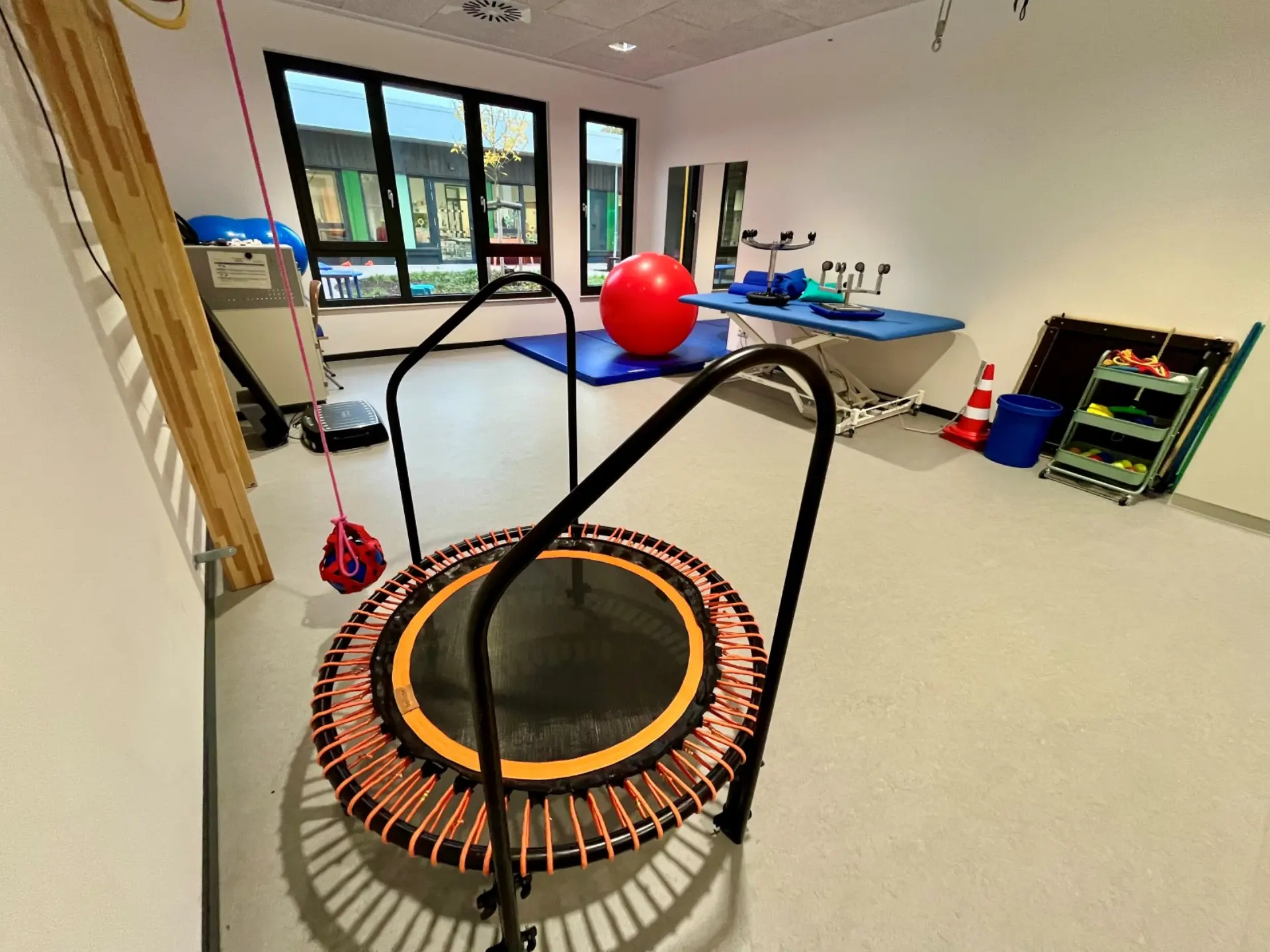 Therapieraum mit Minitrampolin im Vordergrund, Gymnastikball, Behandlungsliege und verschiedenen Sport- und Fitnessgeräten