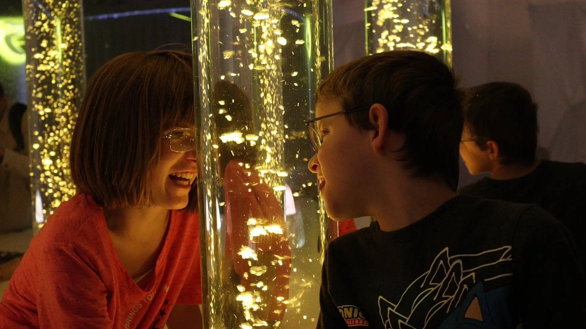Zwei Kinder mit Brille lächeln sich an einer glitzernden Lichtsäule an.