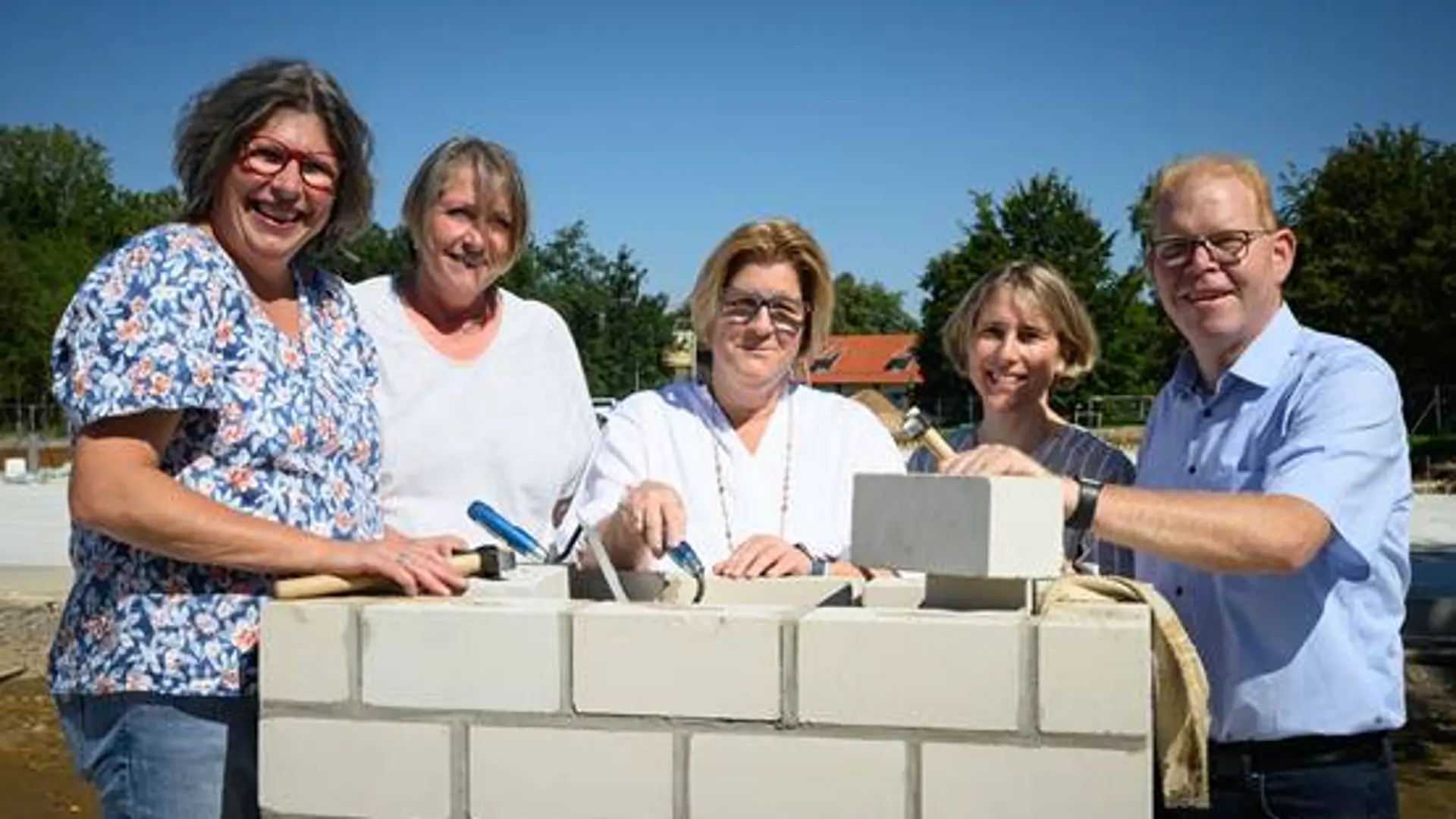 Fünf Personen stehen an einer kleinen gemauerten Wand. Eine hat eine Maurerkelle in der Hand.