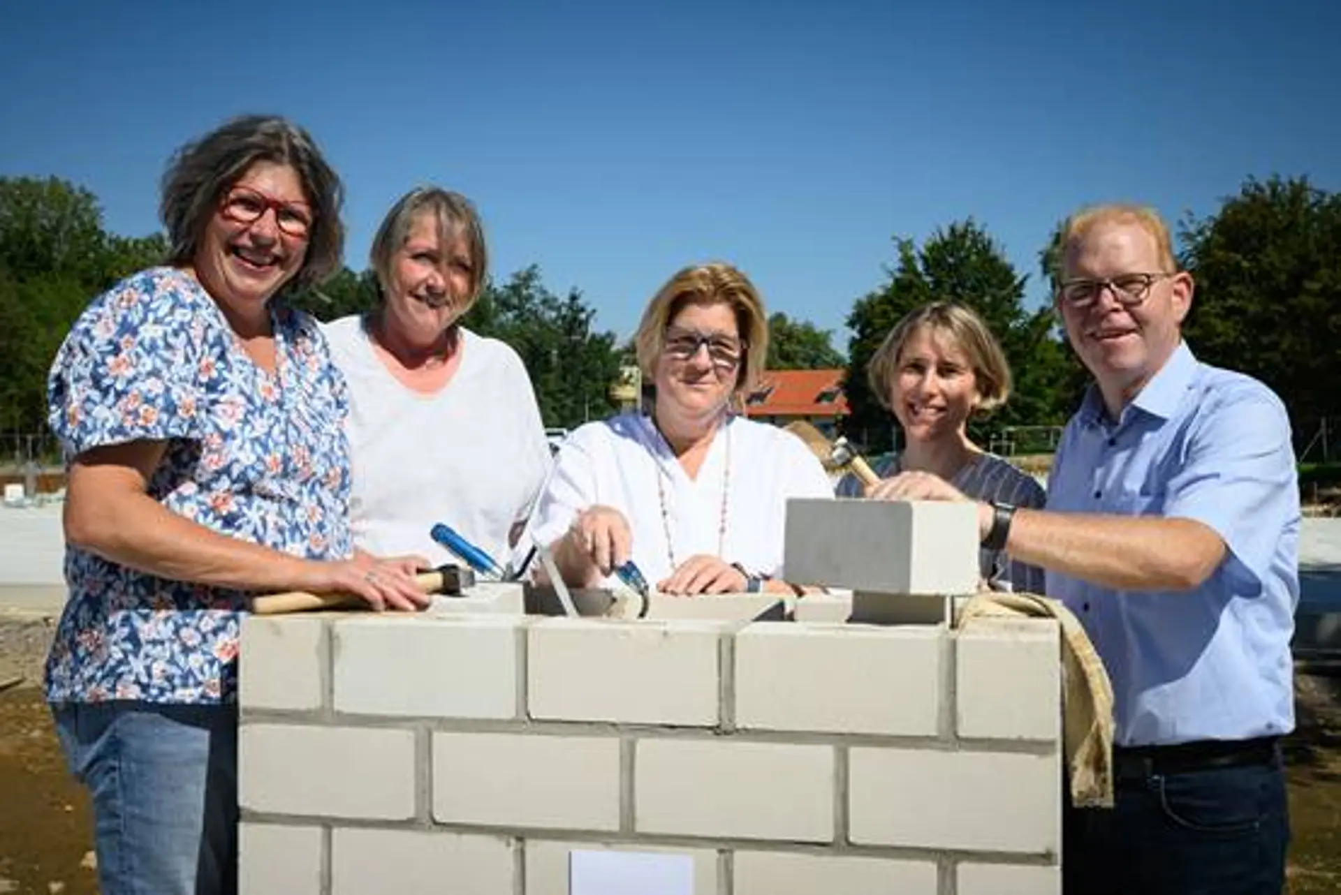 Fünf Personen stehen an einer kleinen gemauerten Wand. Eine hat eine Maurerkelle in der Hand.