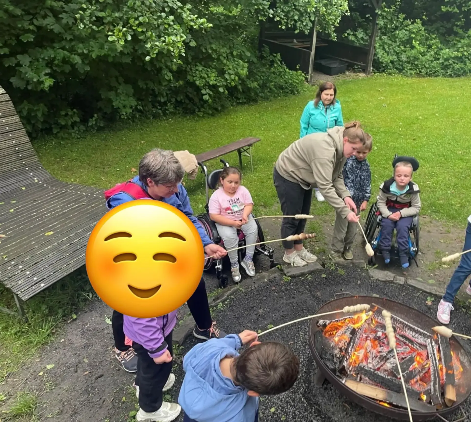 Kinder (zwei von Ihnen im Rollstuhl) und Erwachsene halten Stöcke mit Stockbrot über ein Lagerfeuer in einer Feuerschale 