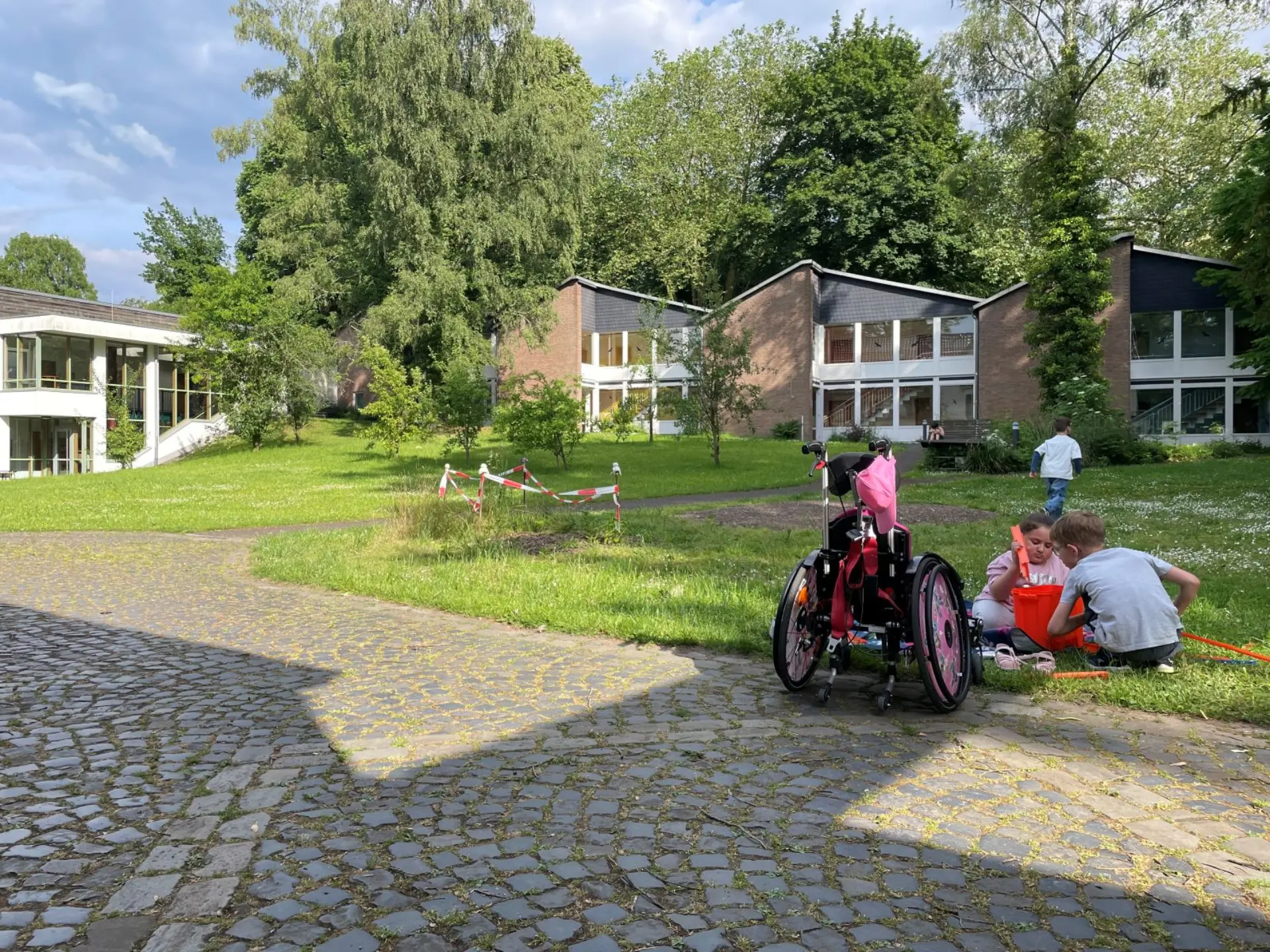 Drei Kinder spielen auf einer Wiese auf einem Gelände mit gepflasterten Wegen und einem Häuserensemble. Neben den Kindern auf dem Weg steht ein Rollstuhl.