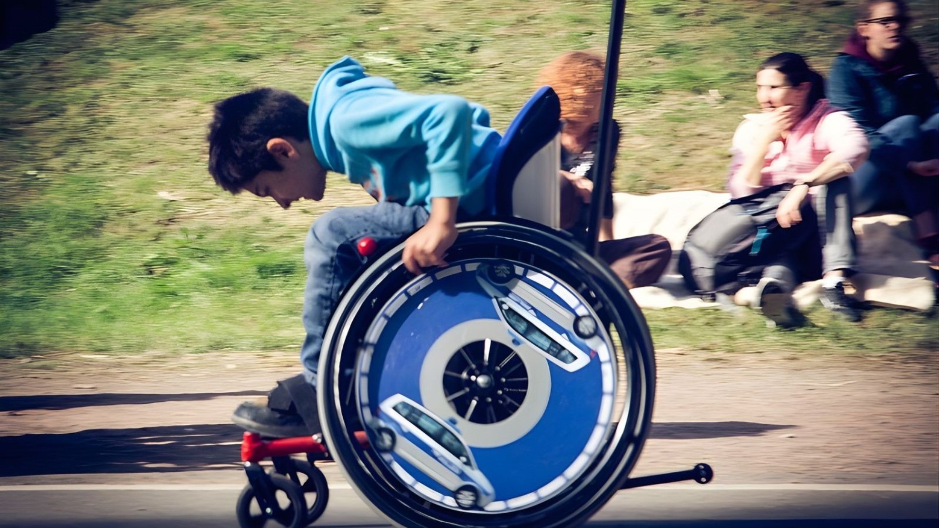 Ein Jugendlicher selbstfahrender Rollstuhlfahrer fährt auf einem asphaltierten Weg von. Er ist von der Seite zu sehen. Das Foto hat eine deutliche Bewegungsunschärfe, die zeigt, dass der Junge schnell unterwegs ist.